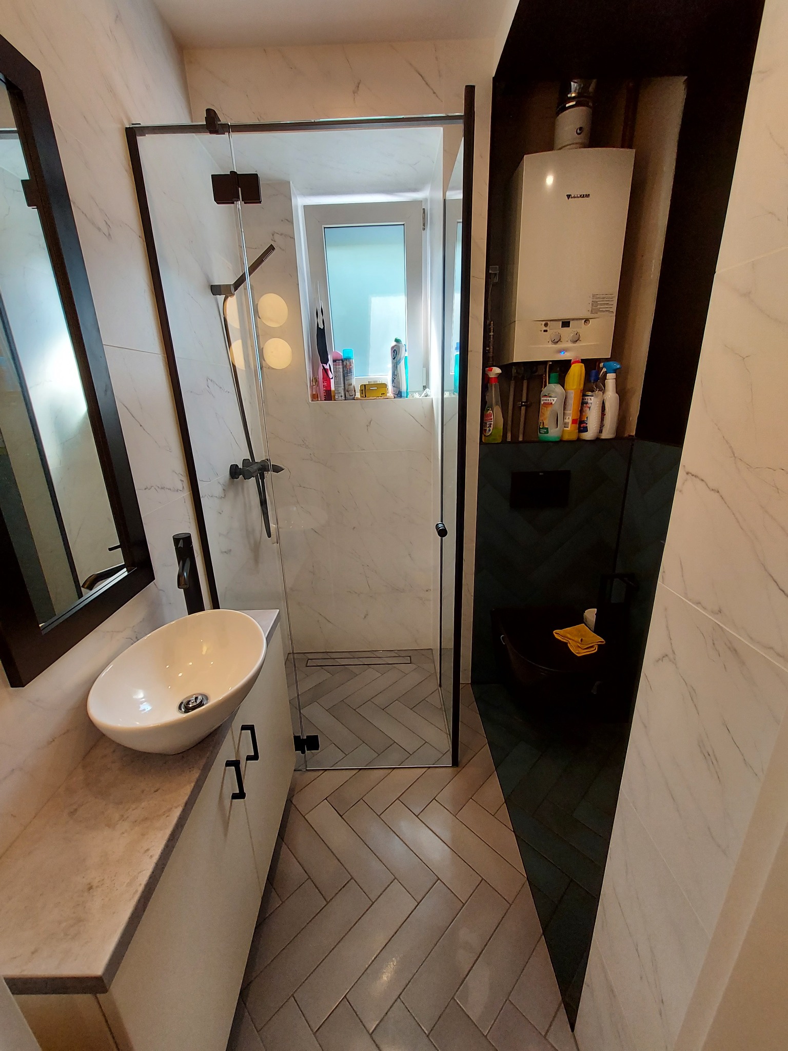 Nowoczesna łazienka z kabiną prysznicową typu walk-in, umywalką nablatową i wiszącym sedesem. Ściany wyłożone płytkami imitującymi marmur, podłoga w jodełkę. Na ścianie nad sedesem zamontowany piec...