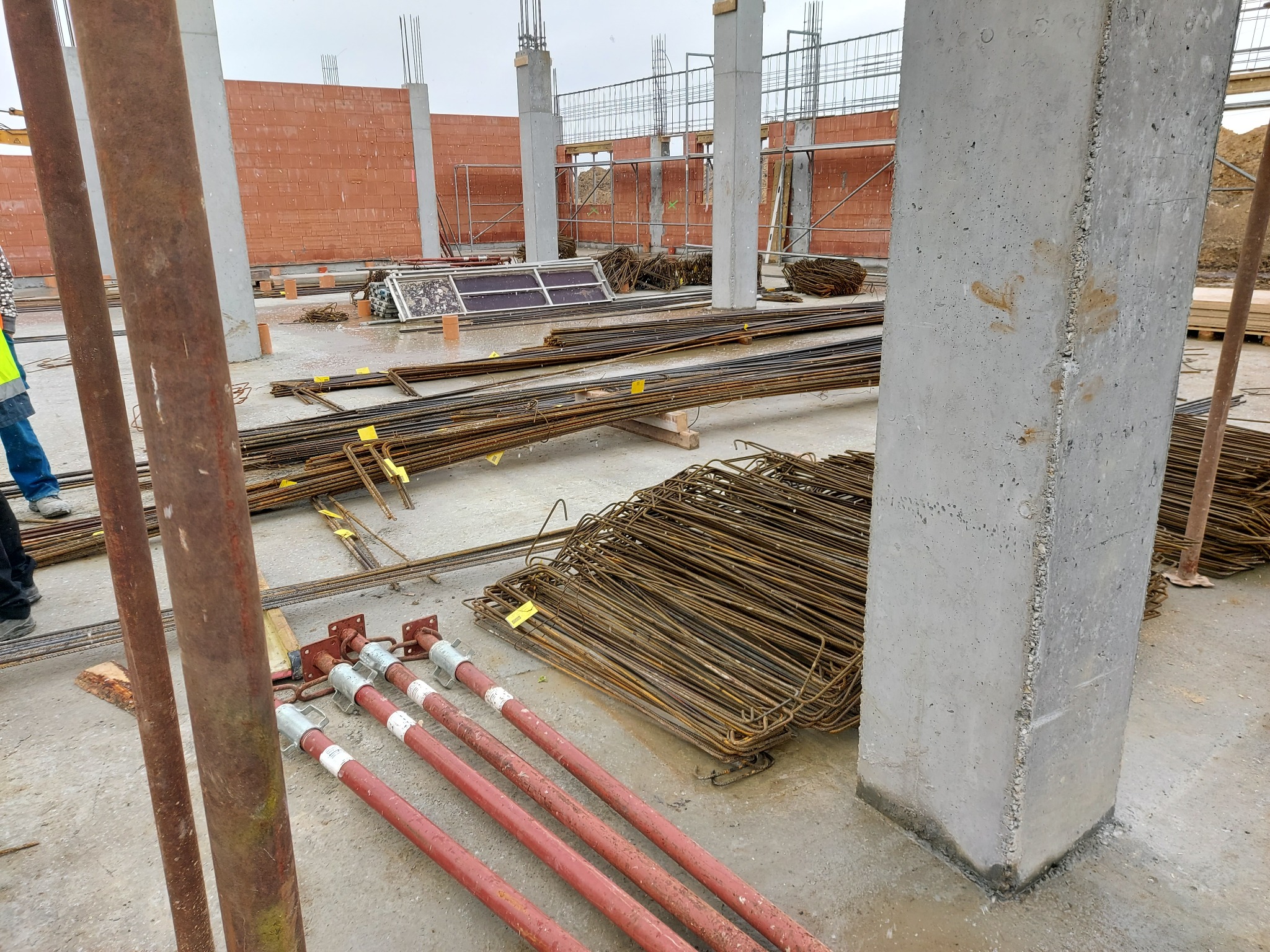 Wiązki zbrojeniowe, stalowe podpory i betonowe słupy na placu budowy, widoczne elementy szalunków i ceglany mur w tle.