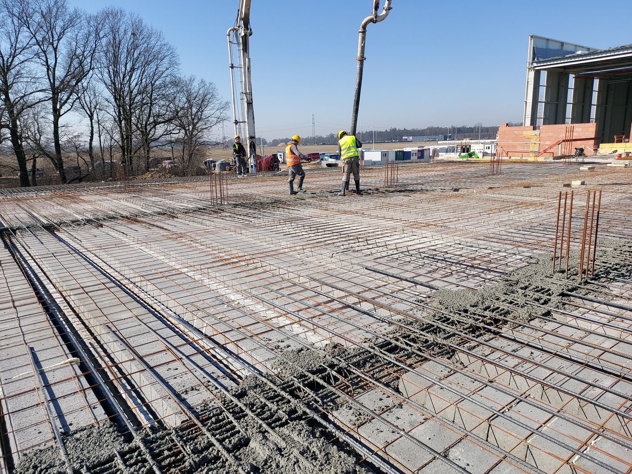 Wylewanie betonu na zbrojoną płytę fundamentową, widoczni pracownicy w odblaskowych kamizelkach i kaskach, w tle krajobraz z drzewami i budynkami.