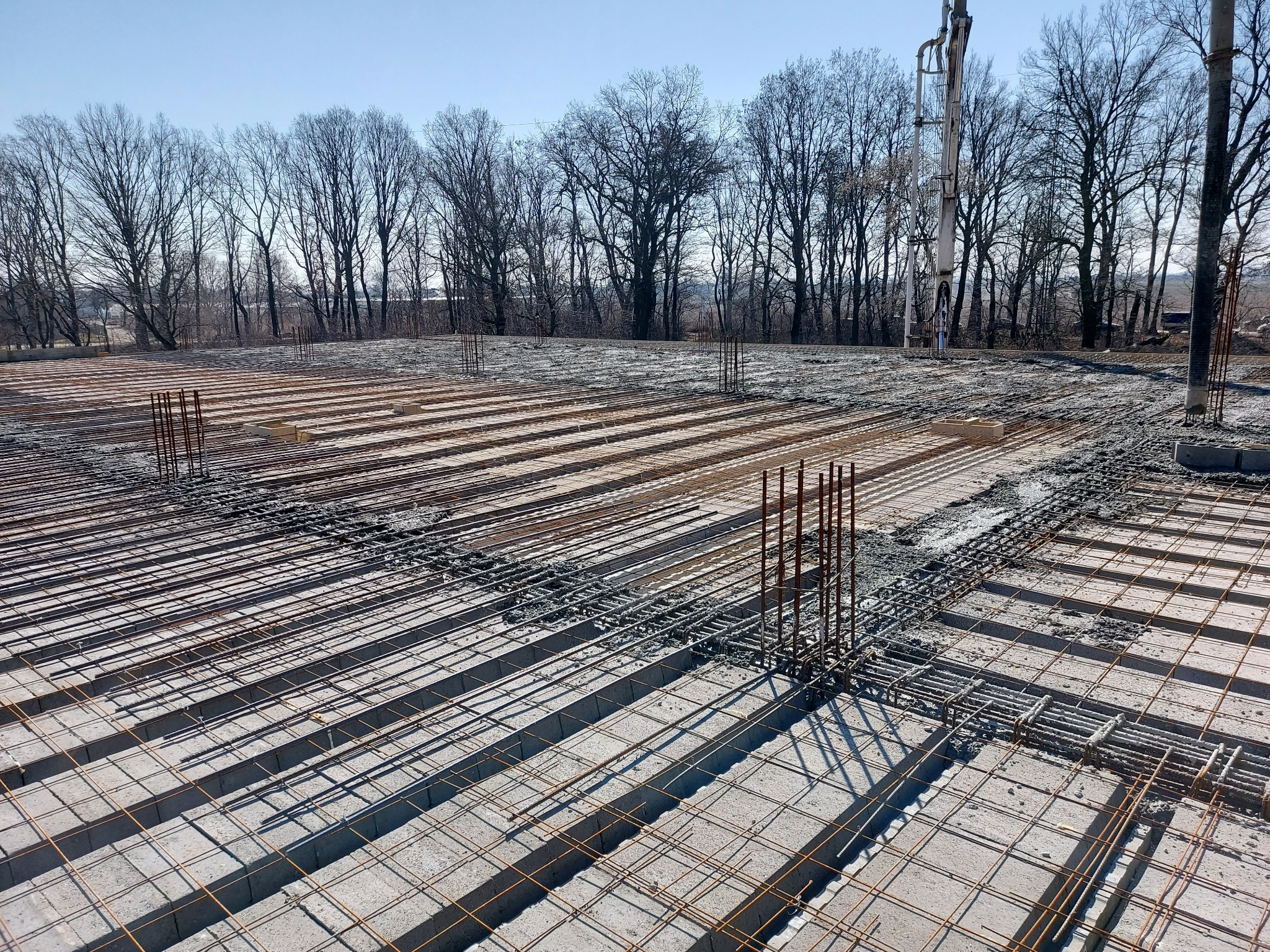 Zbrojenie fundamentów płyty fundamentowej z betonem w trakcie wylewania, widoczna pompa do betonu i drzewa w tle.