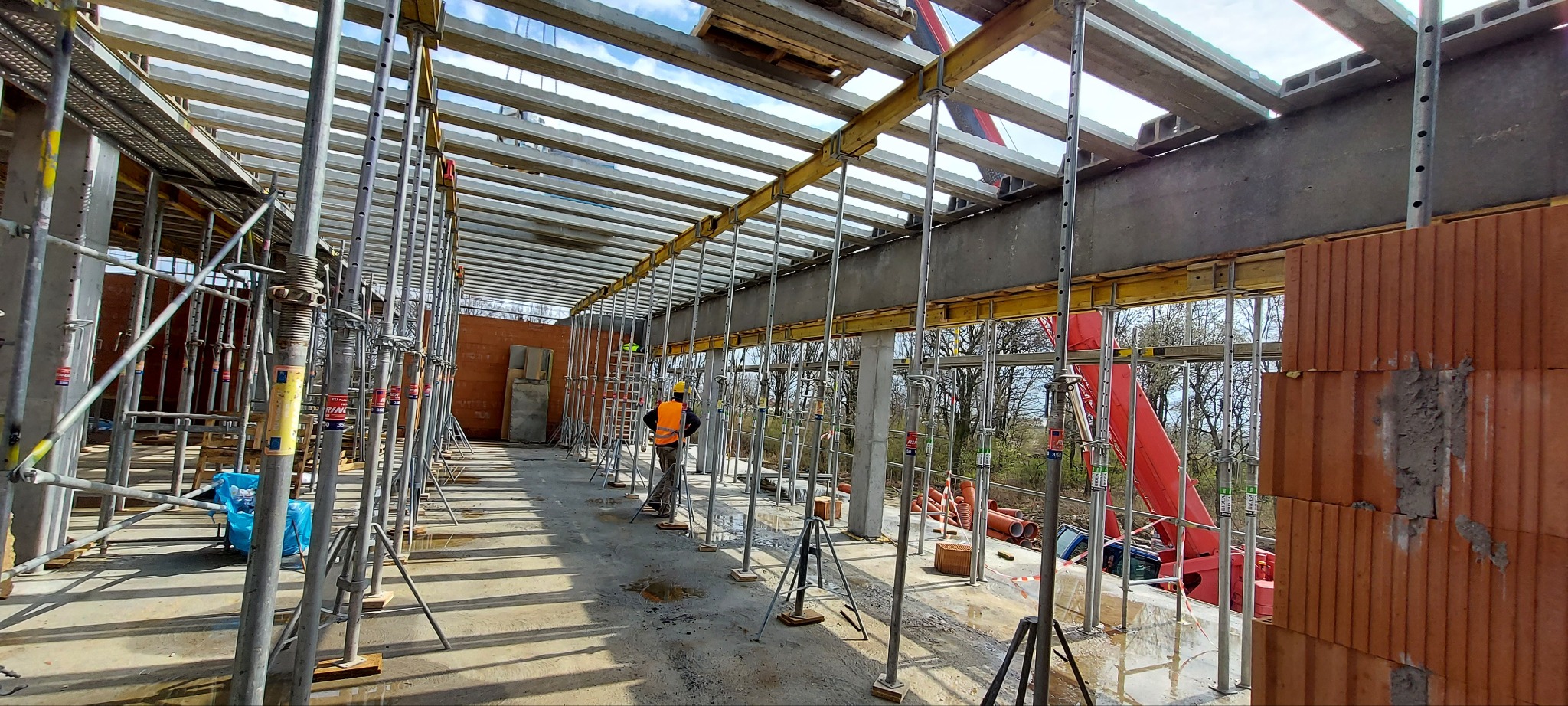 Wnętrze w trakcie budowy z widocznym szalunkiem stropowym, podporami stalowymi, częściowo wymurowanymi ścianami z cegły oraz pracownikiem w pomarańczowej kamizelce i kasku.
