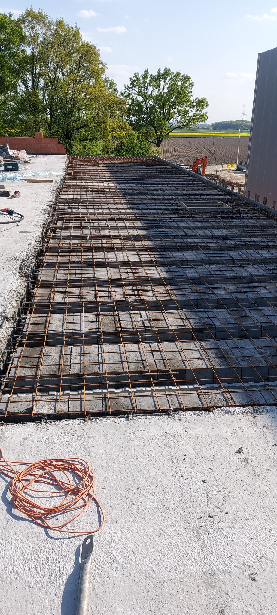 Zbrojenie stropu z pustaków szalunkowych na budowie, widok z góry, w tle drzewa i pole uprawne.