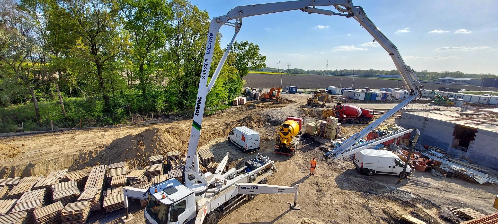 Betonowanie fundamentów budynku: widok z góry na plac budowy z pompą do betonu Schwing, betoniarkami, paletami i pracownikiem w pomarańczowym kombinezonie.
