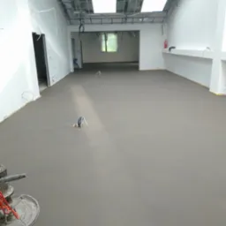 Świeżo wylana, szara posadzka w przestronnym pomieszczeniu z surowym wykończeniem, widoczne przewody elektryczne wystające ze ścian i elementy instalacji pod sufitem.