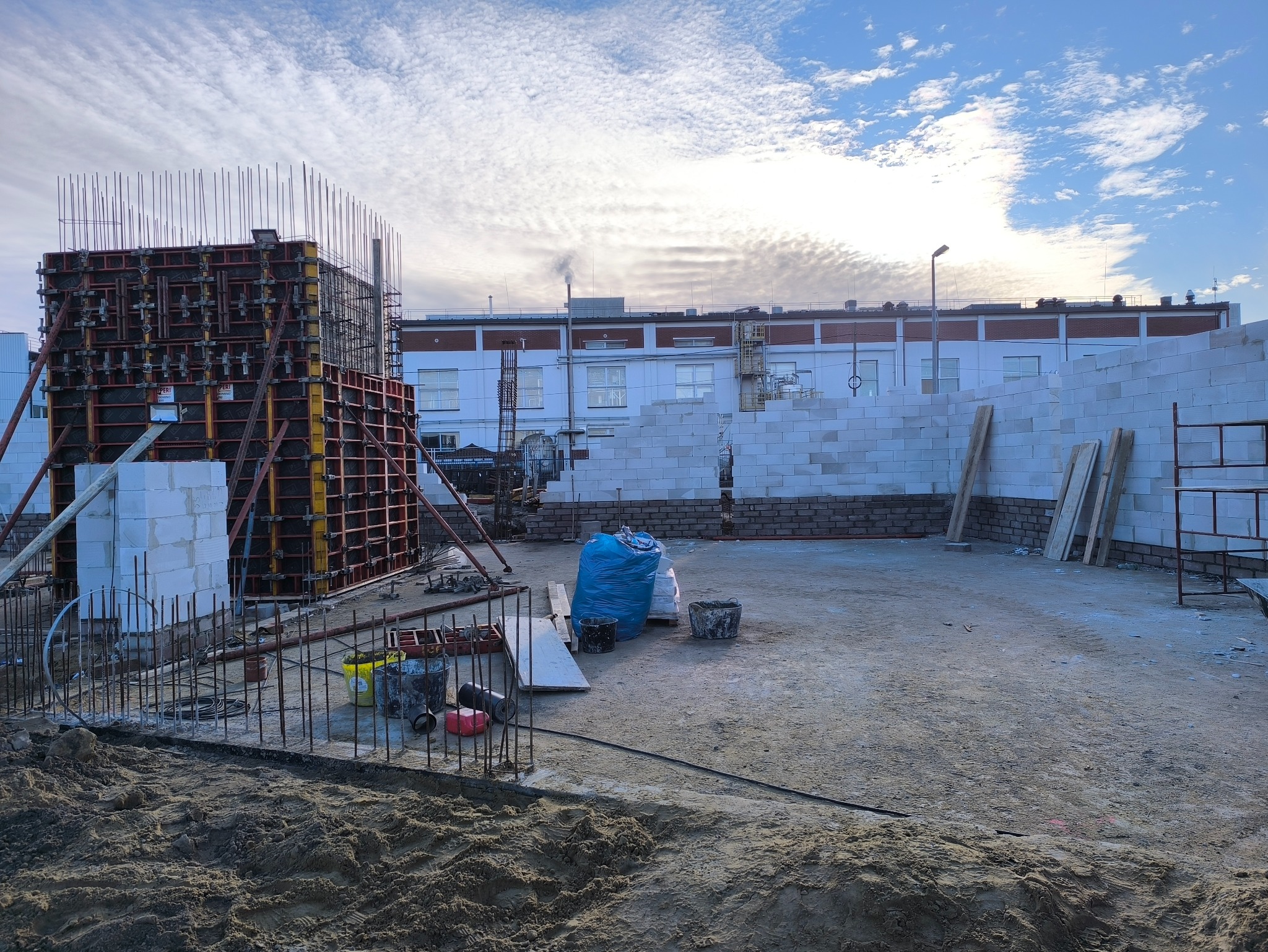 Budowa domu: szalunek zbrojony, ściany z bloczków, materiały budowlane na placu. W tle budynek z kominem i błękitne niebo z chmurami.