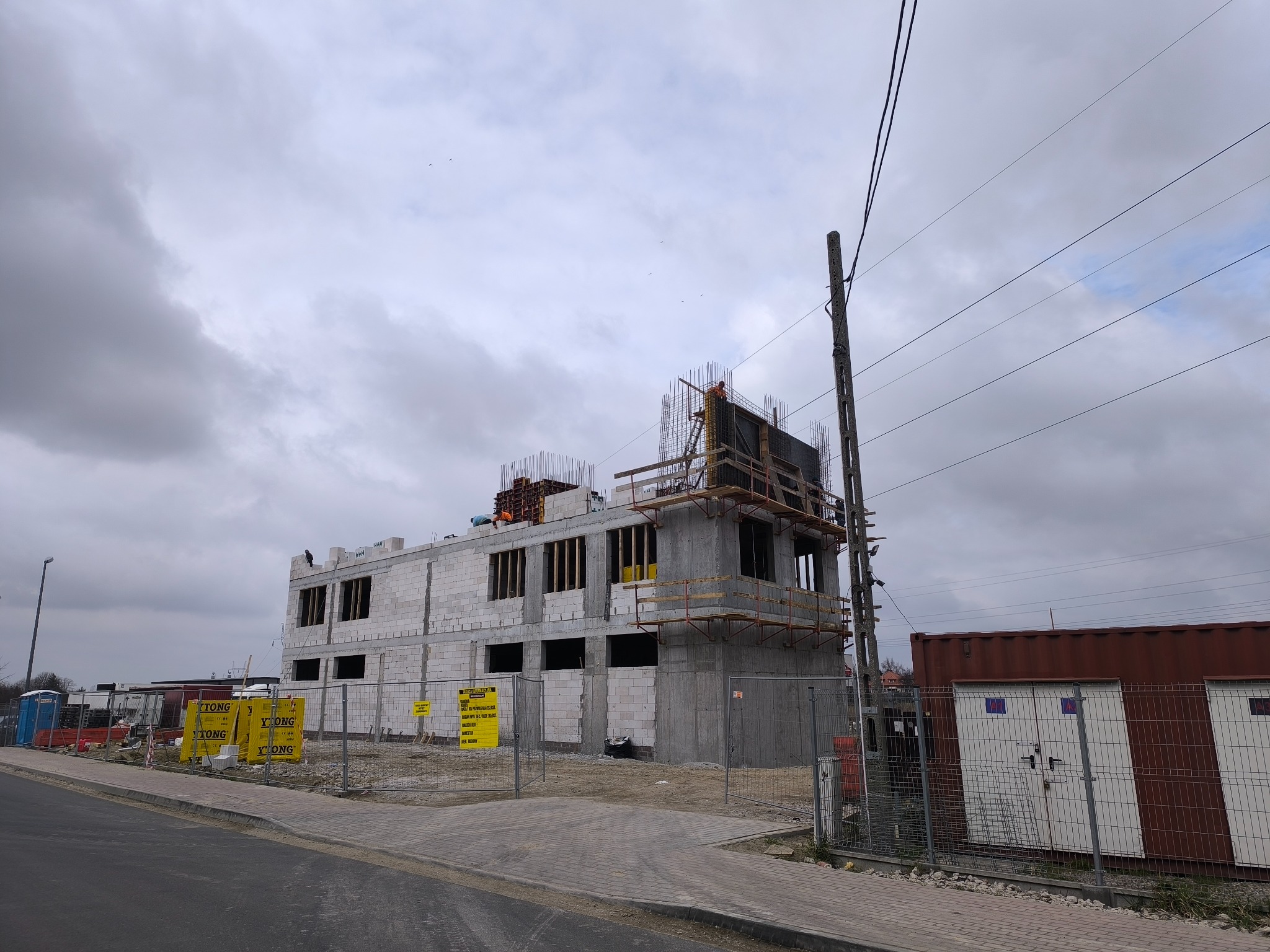 Wznoszony budynek z betonu komórkowego, widoczne zbrojenia i szalunki na dachu. Teren budowy zabezpieczony ogrodzeniem tymczasowym i tablicami informacyjnymi.