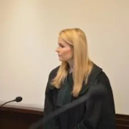 Portret kobiety-adwokata w todze, stojącej za mównicą z mikrofonem, z profilu, na neutralnym tle sali sądowej.