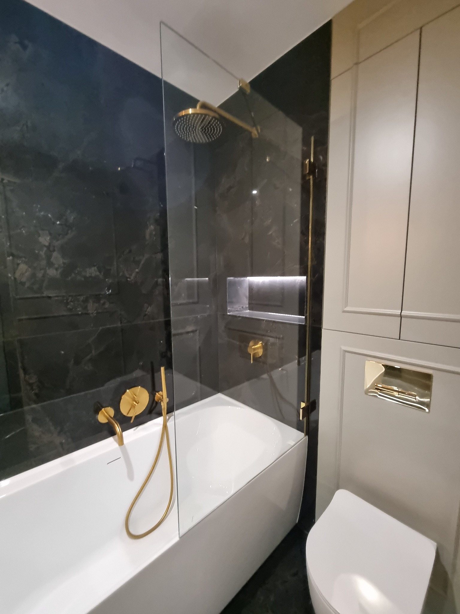 Nowoczesna łazienka: biała wanna z parawanem, złota armatura, czarne marmurowe płytki ścienne z subtelnym frezowaniem i podświetlana wnęka, obok zabudowa meblowa w kolorze kremowym z ukrytą spłuczką.