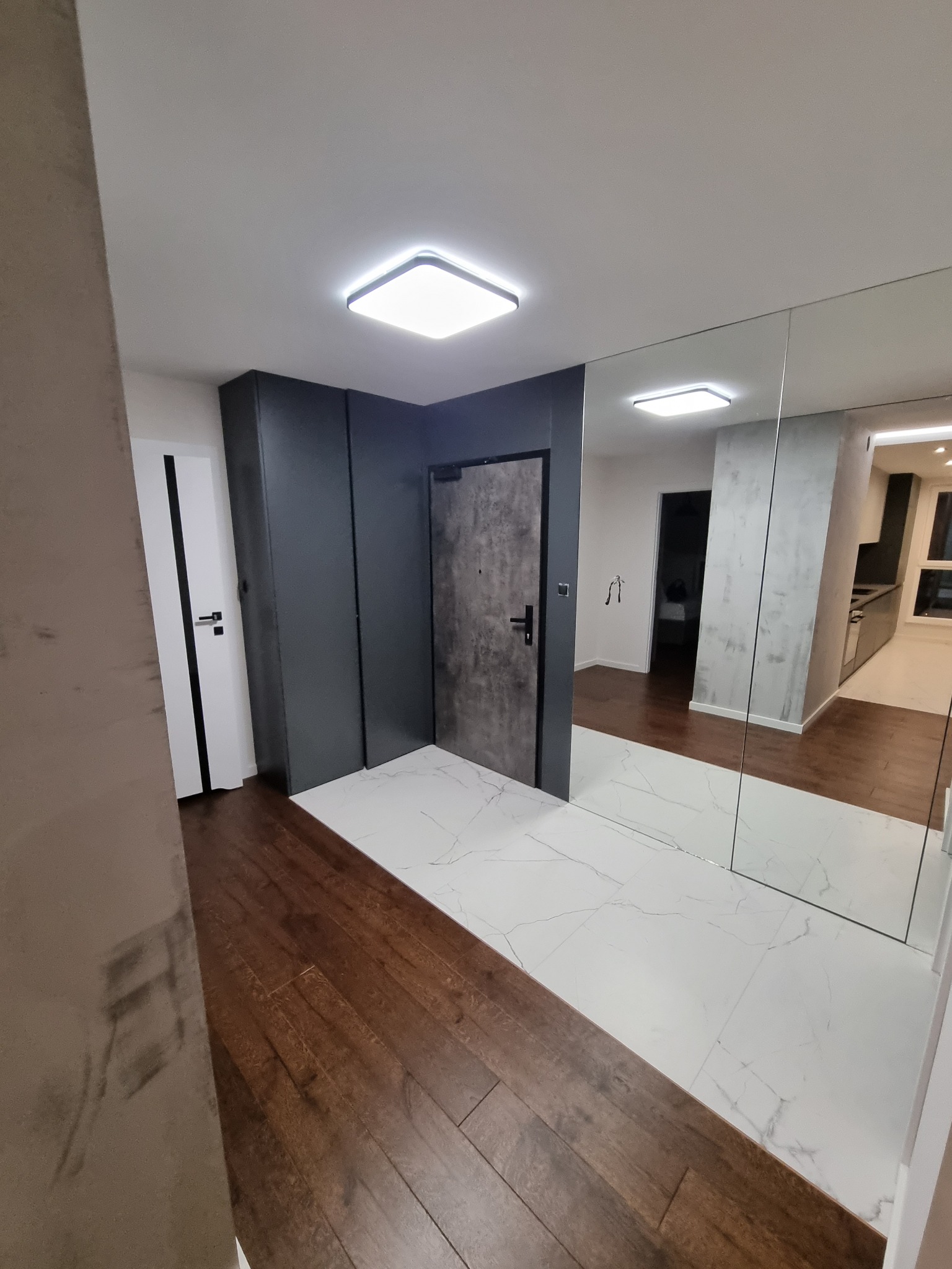 Nowoczesny przedpokój z połączeniem drewnianej podłogi i białych płytek imitujących marmur, szara zabudowa z drzwiami i lustrzaną ścianą odbijającą fragment kuchni.
