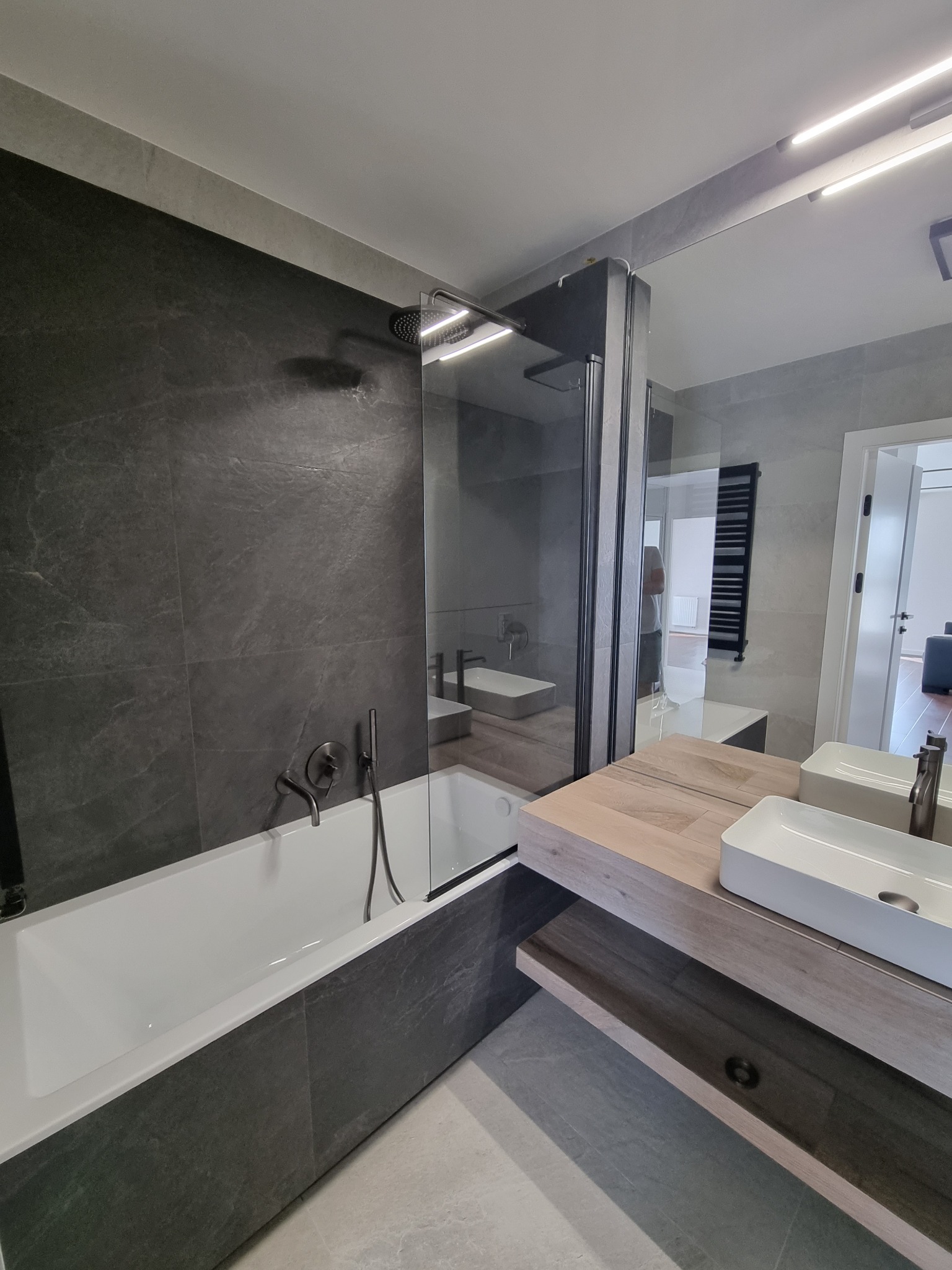 Nowoczesna łazienka z wanną obudowaną ciemnoszarymi płytkami imitującymi kamień, szklanym parawanem nawannowym i umywalką nablatową na drewnianym blacie.