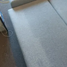 Fragment szarej, modułowej sofy z widocznymi plamami na siedzisku przed czyszczeniem, obok leży żółty wąż odkurzacza piorącego.