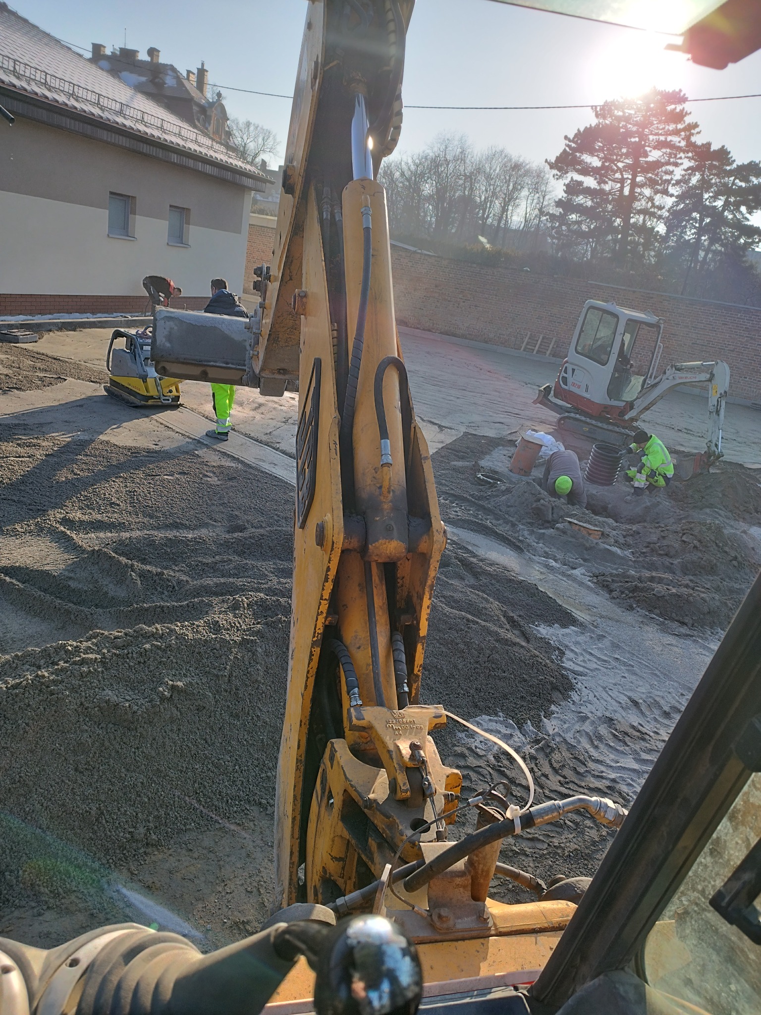 Widok z kabiny koparki na teren budowy, gdzie w oddali widać drugą koparkę, zagęszczarkę oraz robotników w odblaskowych ubraniach pracujących przy instalacji kanalizacyjnej na podsypce piaskowej.