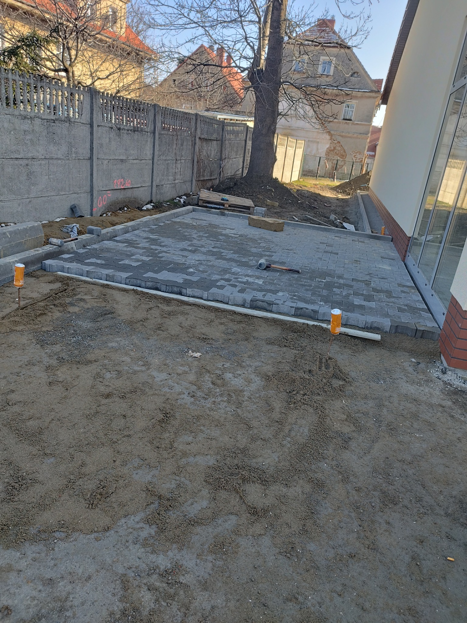 Fragment układanej nawierzchni z szarej kostki brukowej, widoczne narzędzia i elementy wykończeniowe, w tle ogrodzenie i budynek