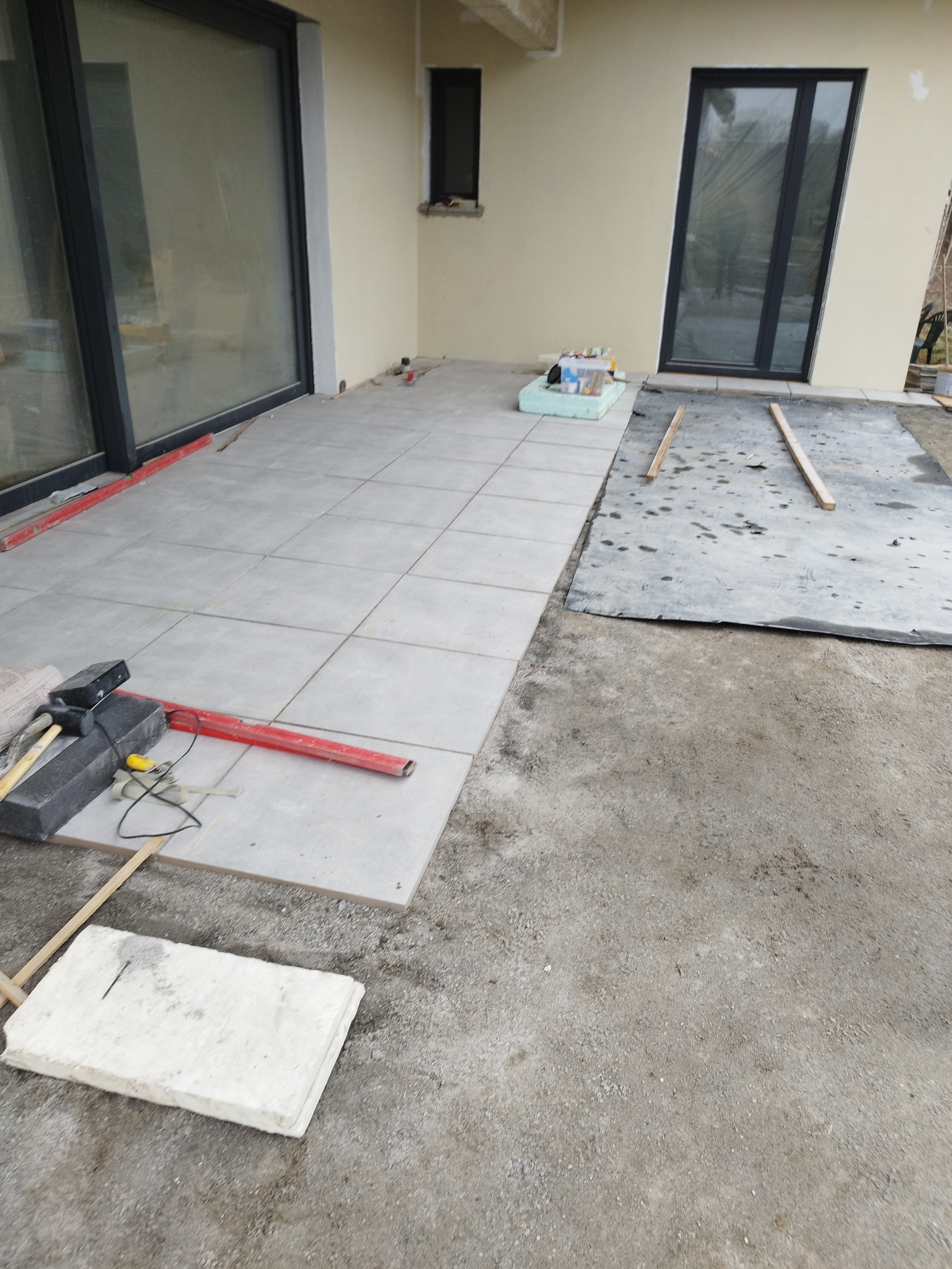 Układanie szarych płyt tarasowych na zewnątrz budynku, widoczne narzędzia brukarskie: poziomica, młotek gumowy, płyty do ułożenia, podłoże częściowo przygotowane, folia ochronna.