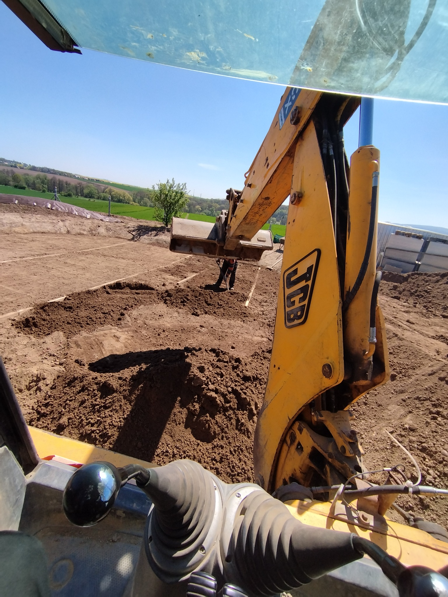 Widok z kabiny koparki JCB na wyrównywany teren pod budowę, z widocznym operatorem i świeżo przekopaną ziemią, w tle zielone pola i błękitne niebo.