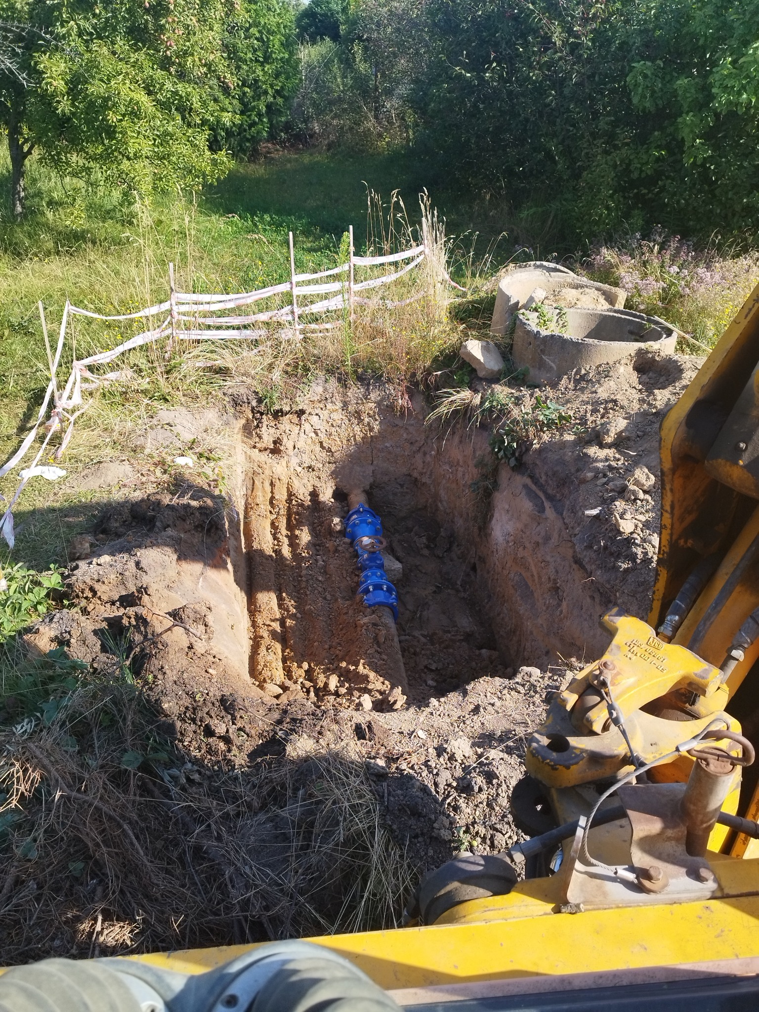 Głęboki wykop w ziemi z widocznymi niebieskimi rurami łączonymi złączkami, fragment żółtej koparki w prawym dolnym rogu kadru, teren zabezpieczony białą taśmą, w tle zieleń drzew