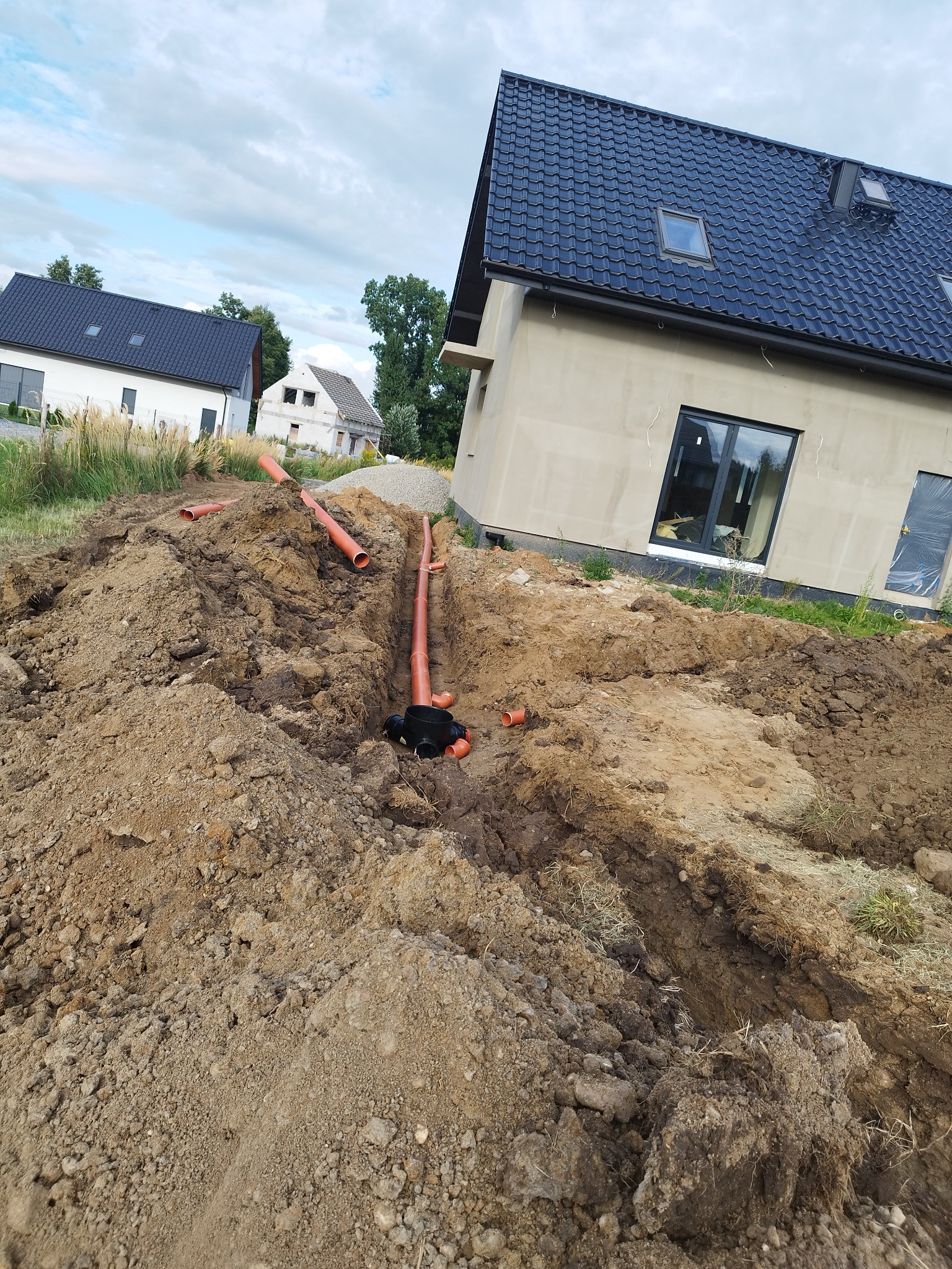 Wykopany rów z ułożonymi rurami kanalizacyjnymi z tworzywa sztucznego na tle nowo wybudowanego domu z czarnym dachem i szarymi ścianami.