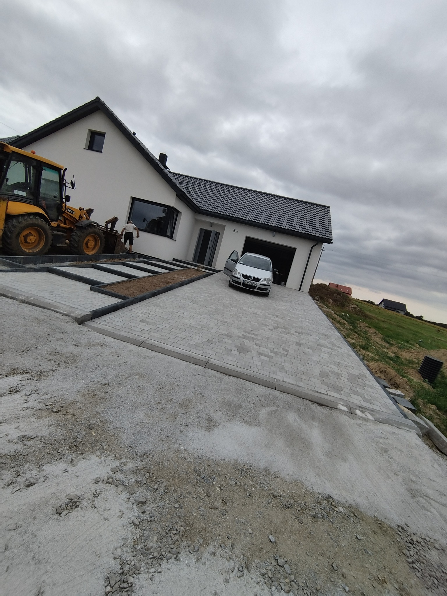Układanie kostki brukowej na podjeździe w Świdnicy, widoczny żółty traktor i biały dom z garażem, etap prac ziemnych.