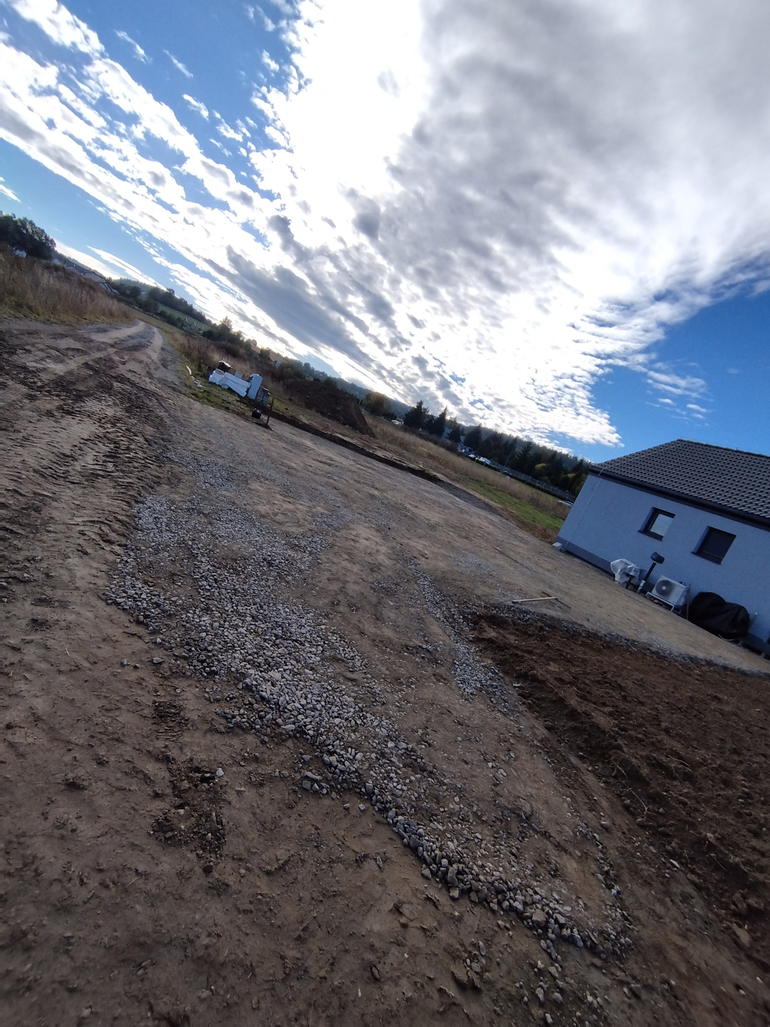 Przygotowany teren pod budowę domu z nasypaną warstwą gruzu, widoczne ślady opon i nowo wybudowany dom w tle pod pochmurnym niebem.