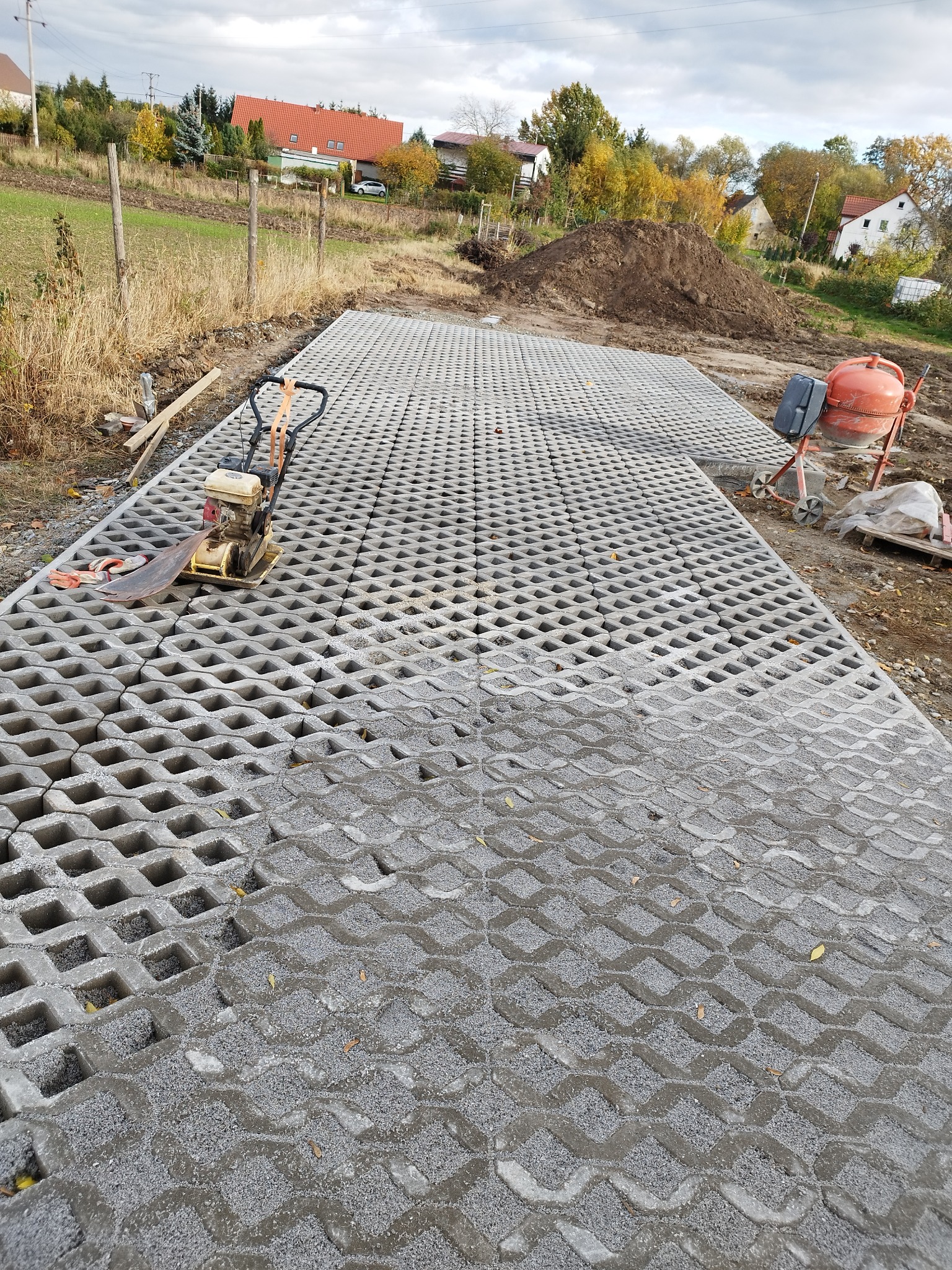 Ułożona nawierzchnia z ażurowych płyt betonowych wypełnionych szarym kruszywem, widoczny zagęszczarka i betoniarka, w tle krajobraz wiejski z domami i stertą ziemi.