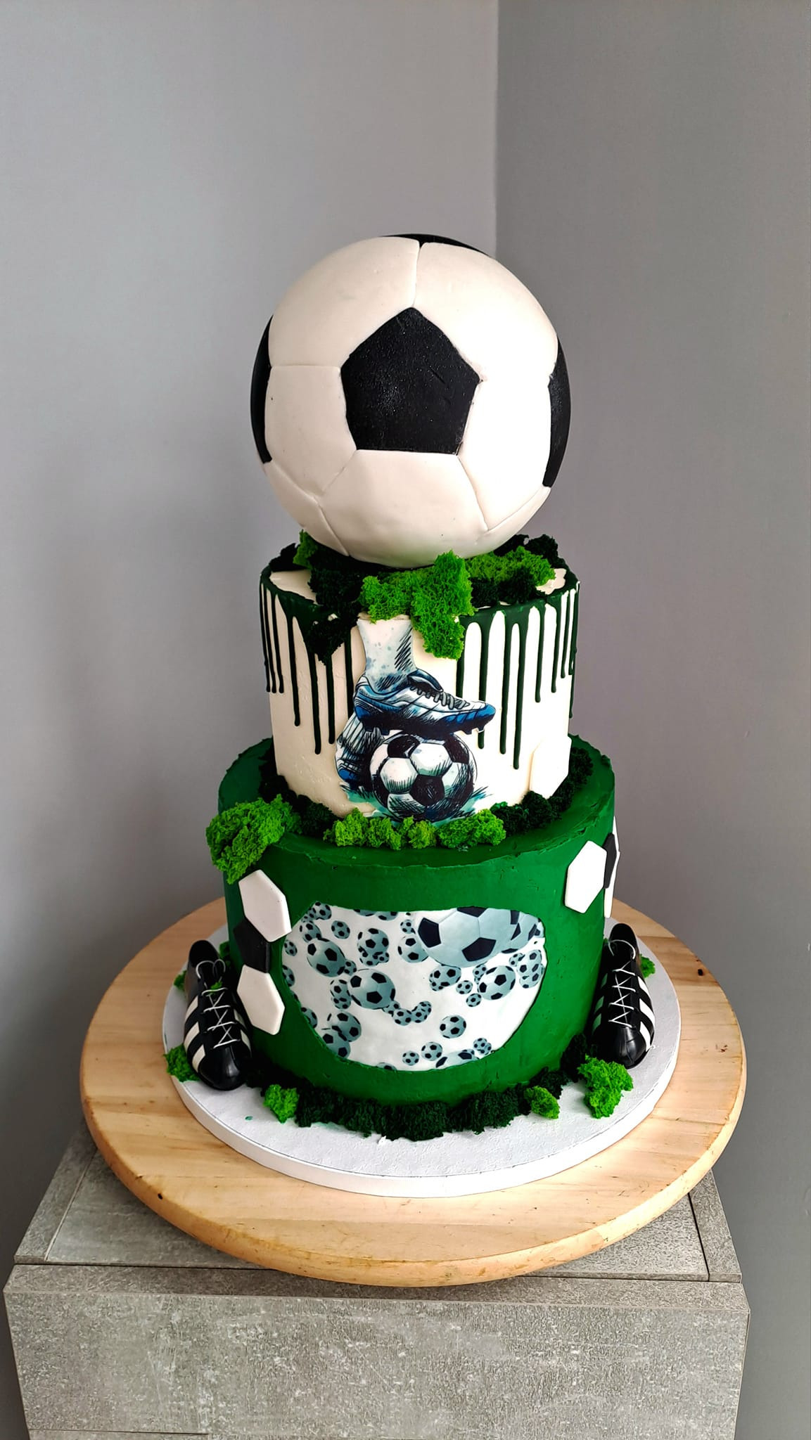 Trzypiętrowy tort tematyczny piłkarski, z dużą jadalną piłką na szczycie, zielonym i białym lukrem, dekoracjami z mchu cukrowego i małymi butami piłkarskimi z lukru u podstawy.