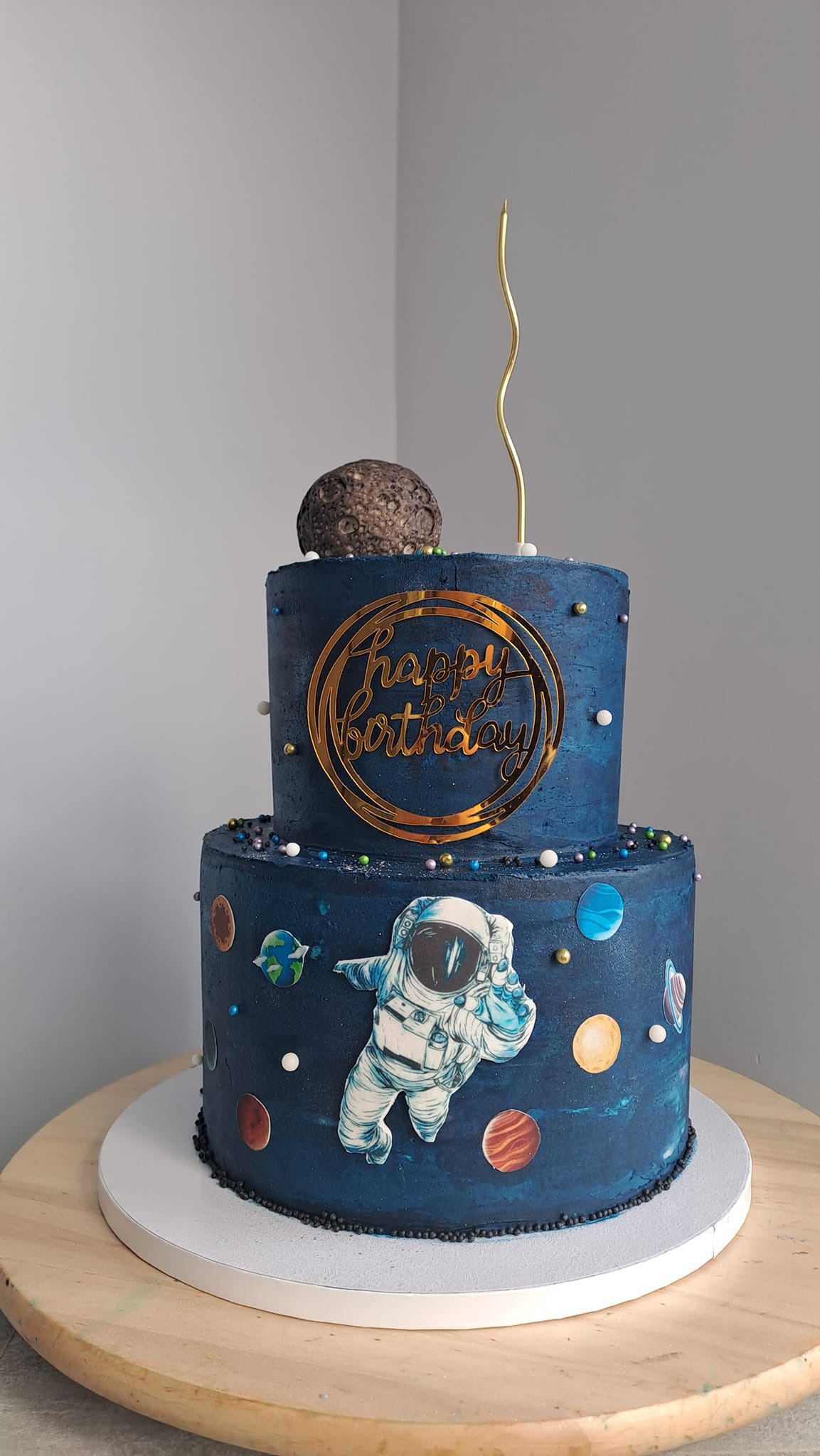 Dwupoziomowy tort urodzinowy w kosmicznym stylu: ciemnoniebieski krem, dekoracje z planet, figurka astronauty, topper 'Happy Birthday' i imitacja księżyca na szczycie.