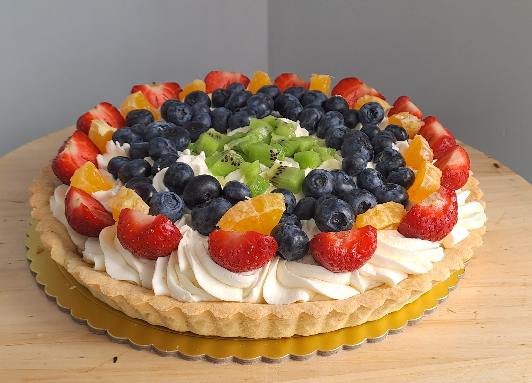 Okrągły tort owocowy z kremem, udekorowany świeżymi truskawkami, borówkami, pomarańczami i kiwi, umieszczony na złotej podkładce.