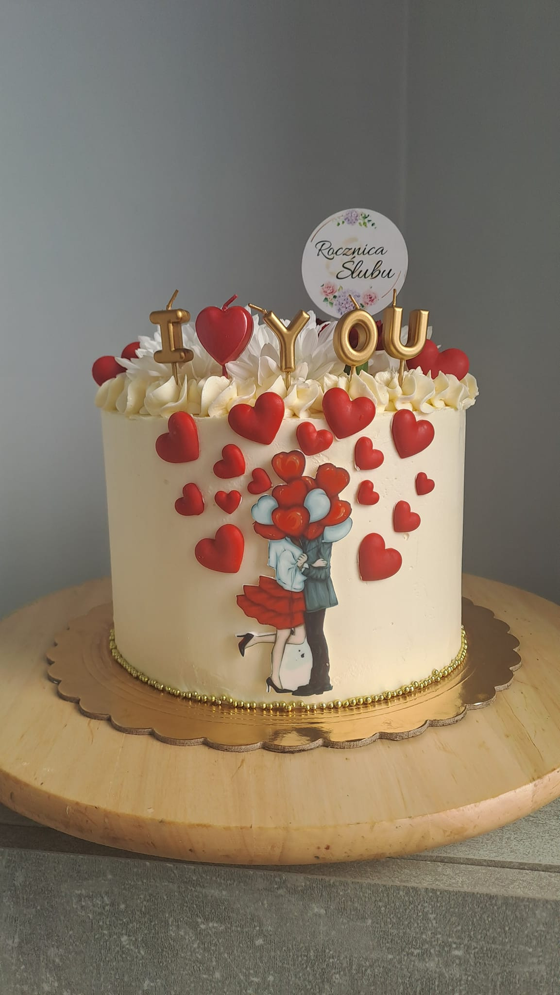 Tort rocznicowy z kremem, dekoracjami z czerwonych serc, topperem 'I <3 YOU' i wizerunkiem całującej się pary z balonami w kształcie serc.