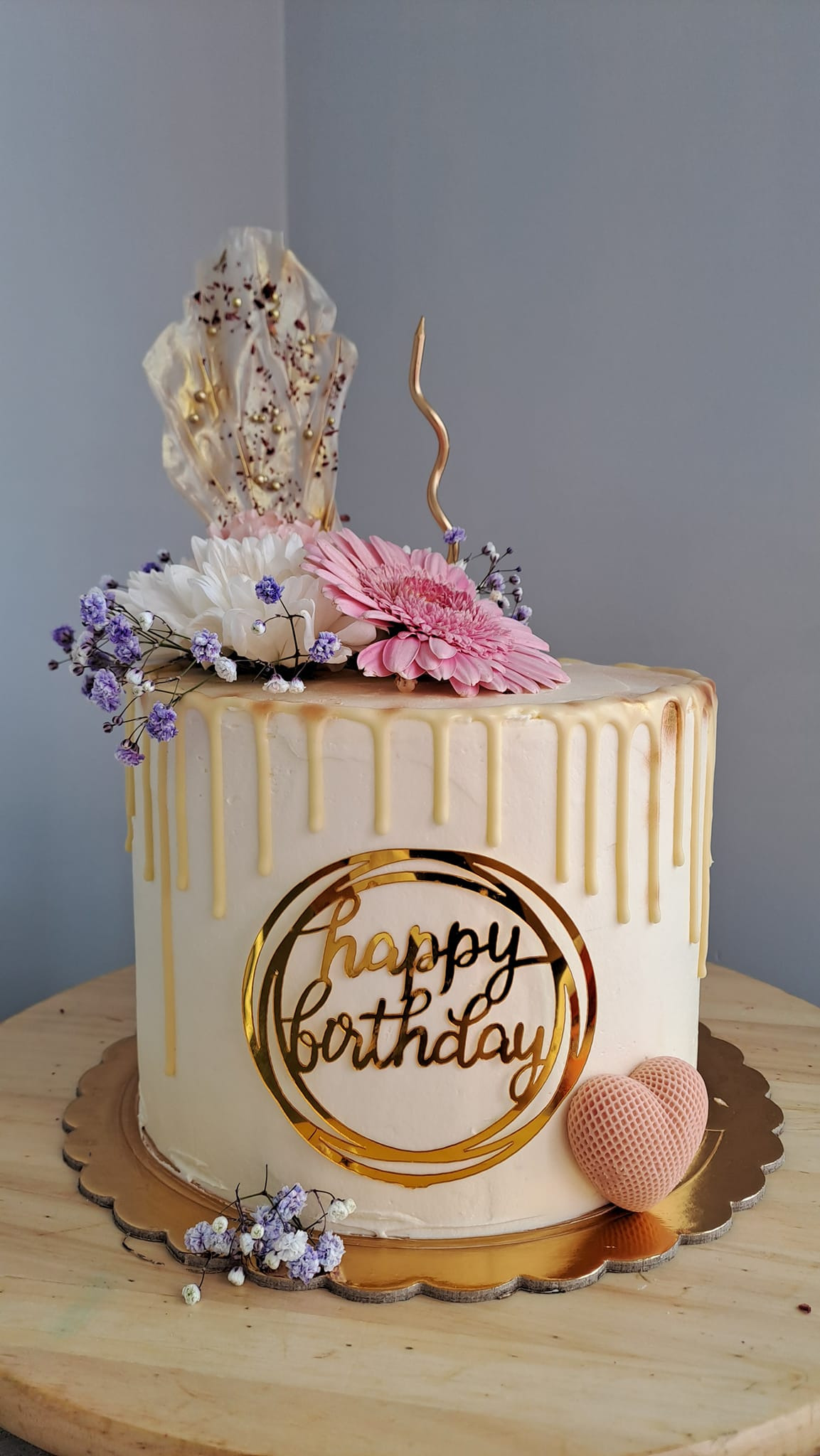 Elegancki tort urodzinowy z białym kremem, polewą, dekoracją z kwiatów, złotym napisem 'Happy Birthday' i ozdobnym wachlarzem z jadalnego papieru.