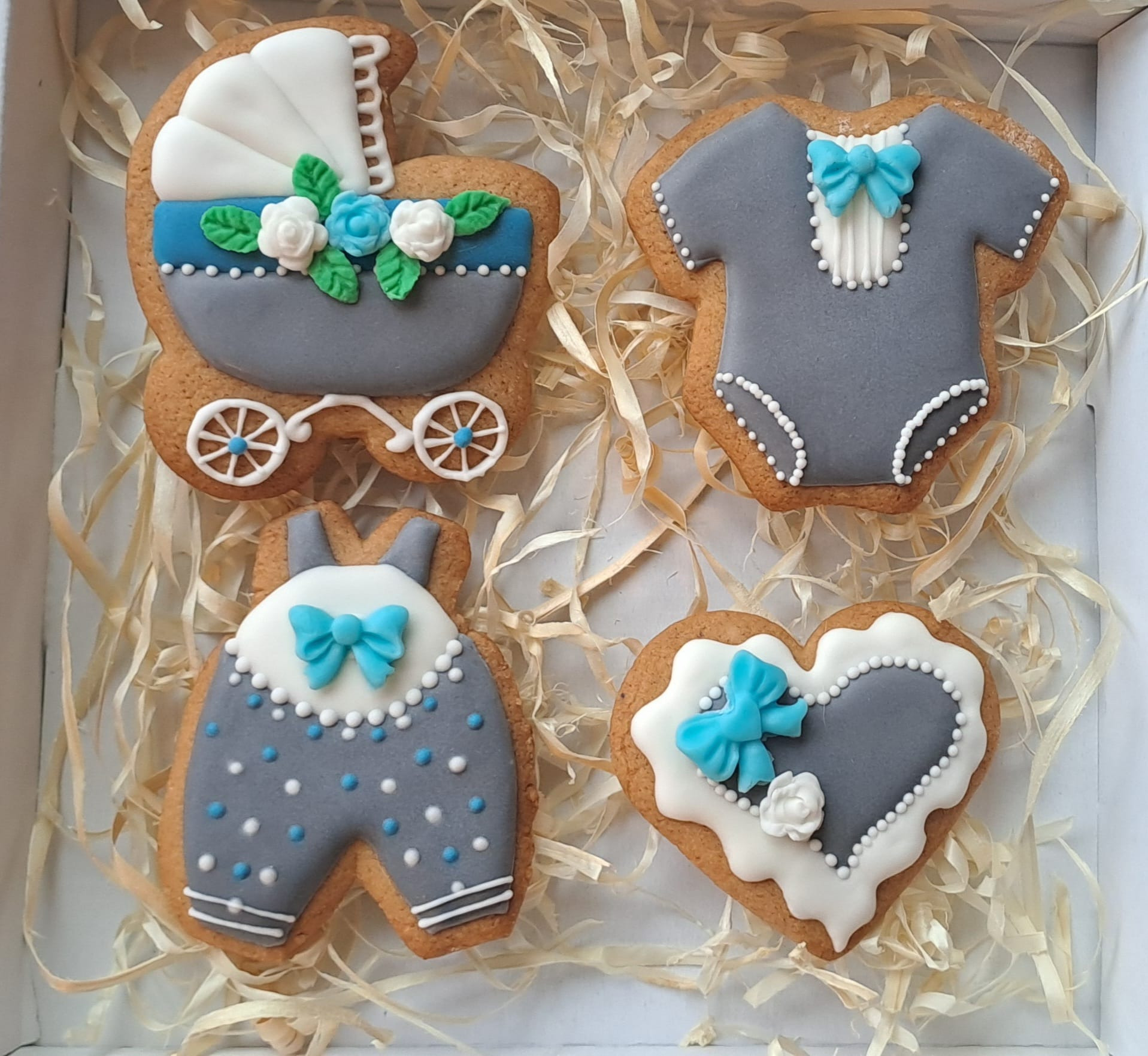 Ozdobne pierniczki w kształcie wózka dziecięcego, niemowlęcego body, śpioszków i serca, udekorowane lukrem w odcieniach szarości, bieli i błękitu, umieszczone w pudełku wypełnionym siankiem.