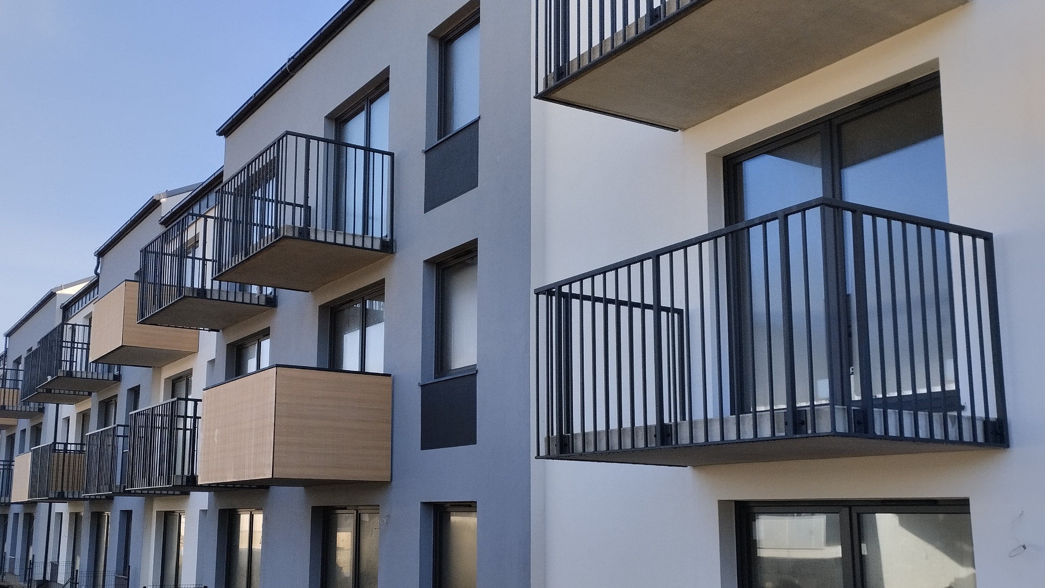 Nowoczesny budynek wielorodzinny z balkonami o czarnych, metalowych balustradach i częściowo zabudowanymi ściankami w kolorze jasnego drewna, widziany pod kątem z dołu w słoneczny dzień.