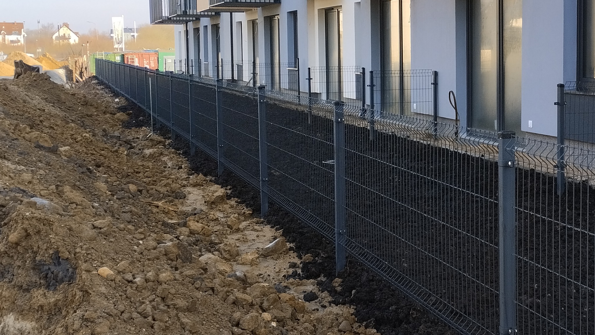 Nowoczesne ogrodzenie panelowe z ciemnoszarej siatki, zabezpieczające teren budowy w Szczecinie, z widocznymi świeżo posadzonymi roślinami u podstawy i budynkiem mieszkalnym w tle.