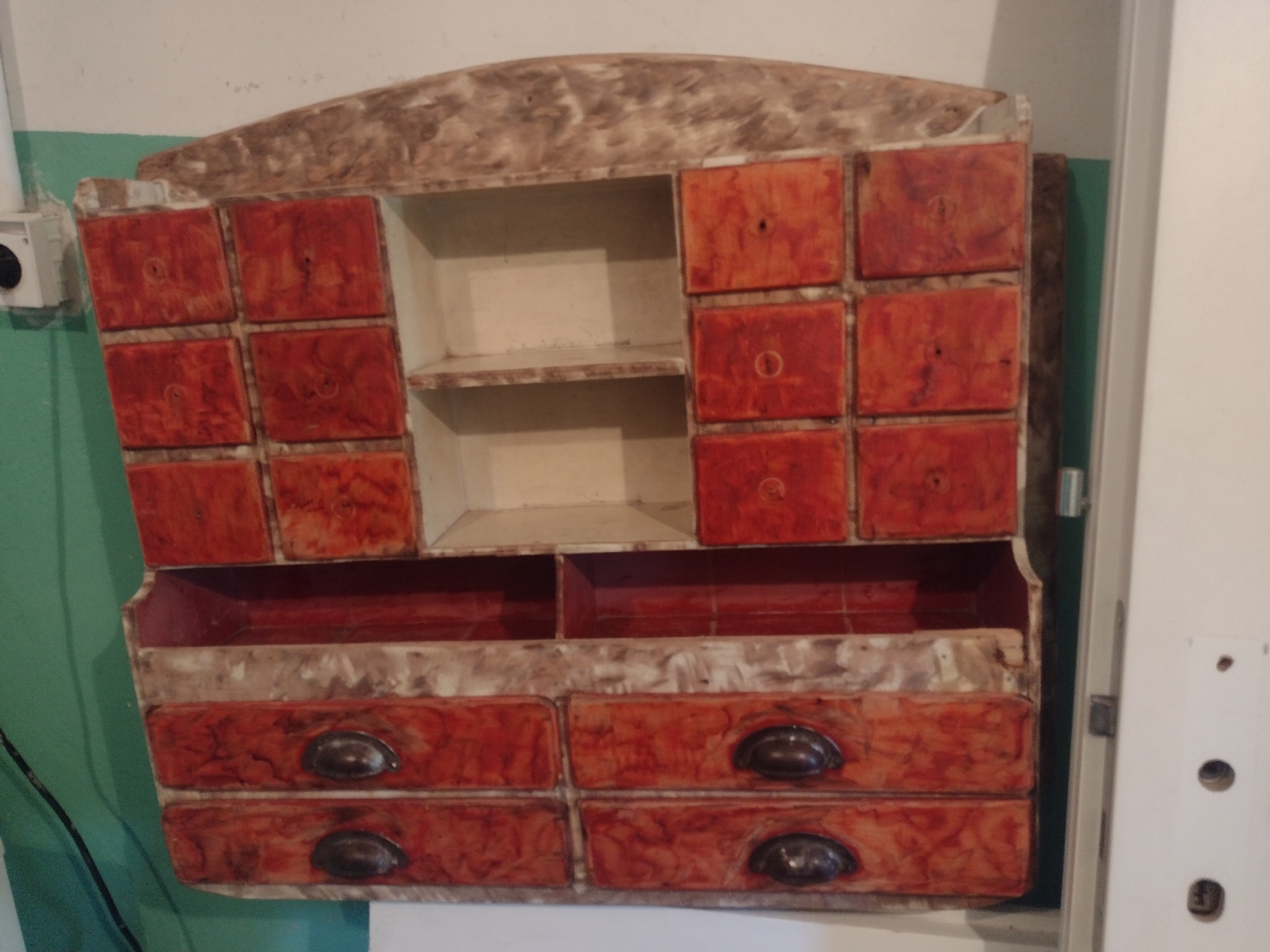 Antyczna, drewniana szafka z szufladami i półkami, z widocznymi śladami użytkowania i odbarwieniami farby, zawieszona na zielonej ścianie.