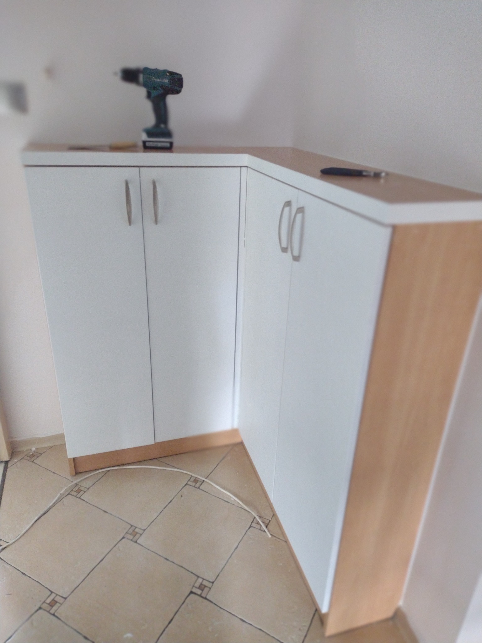 Białe, narożne szafki z drewnianymi akcentami i srebrnymi uchwytami, na blacie wiertarka akumulatorowa i ołówek, na podłodze płytki i kabel.