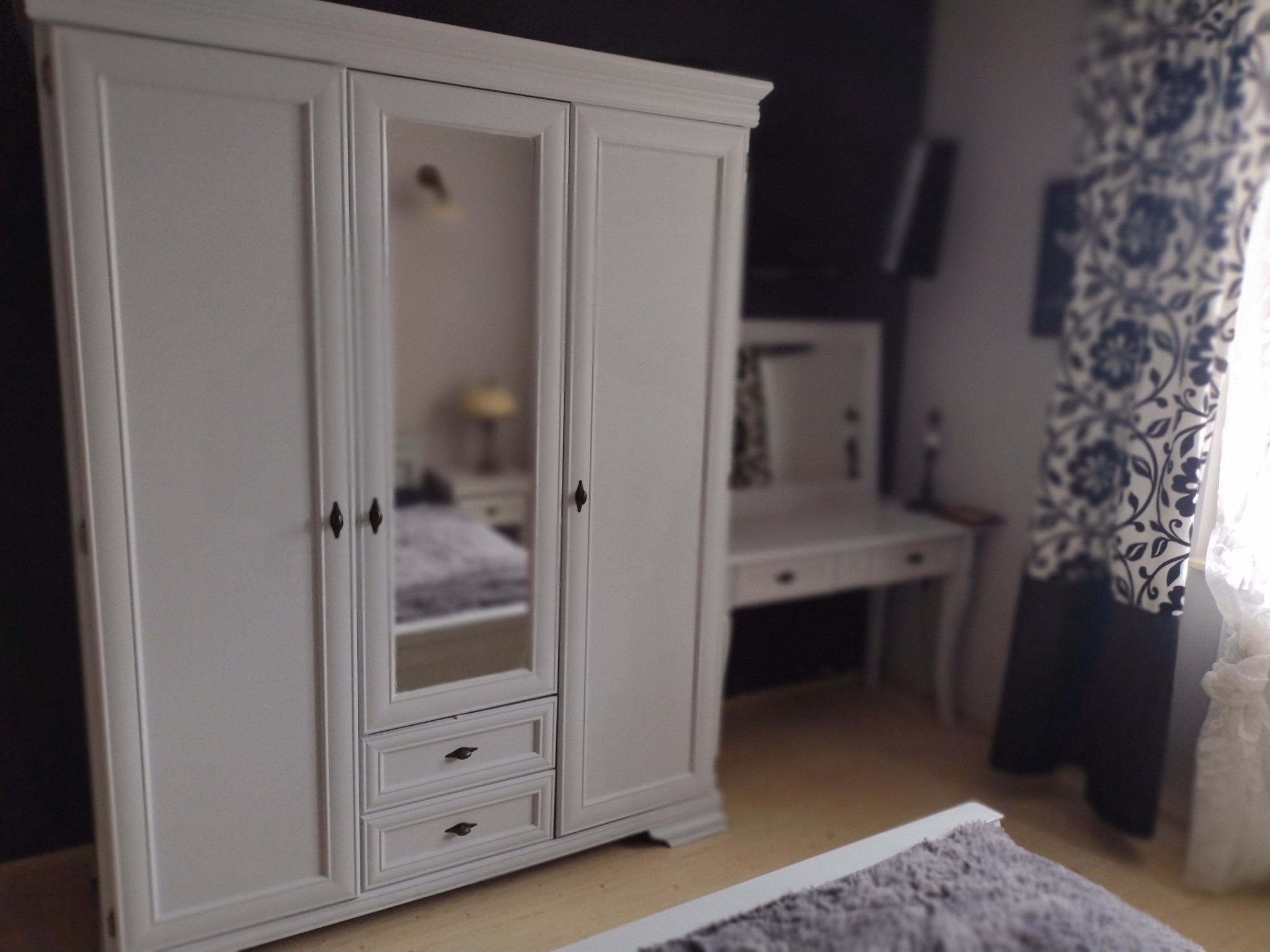 Biała, trzydrzwiowa szafa z lustrem w środkowych drzwiach, uzupełniona o dwie szuflady u dołu, w aranżacji sypialni z toaletką i wzorzystymi zasłonami.