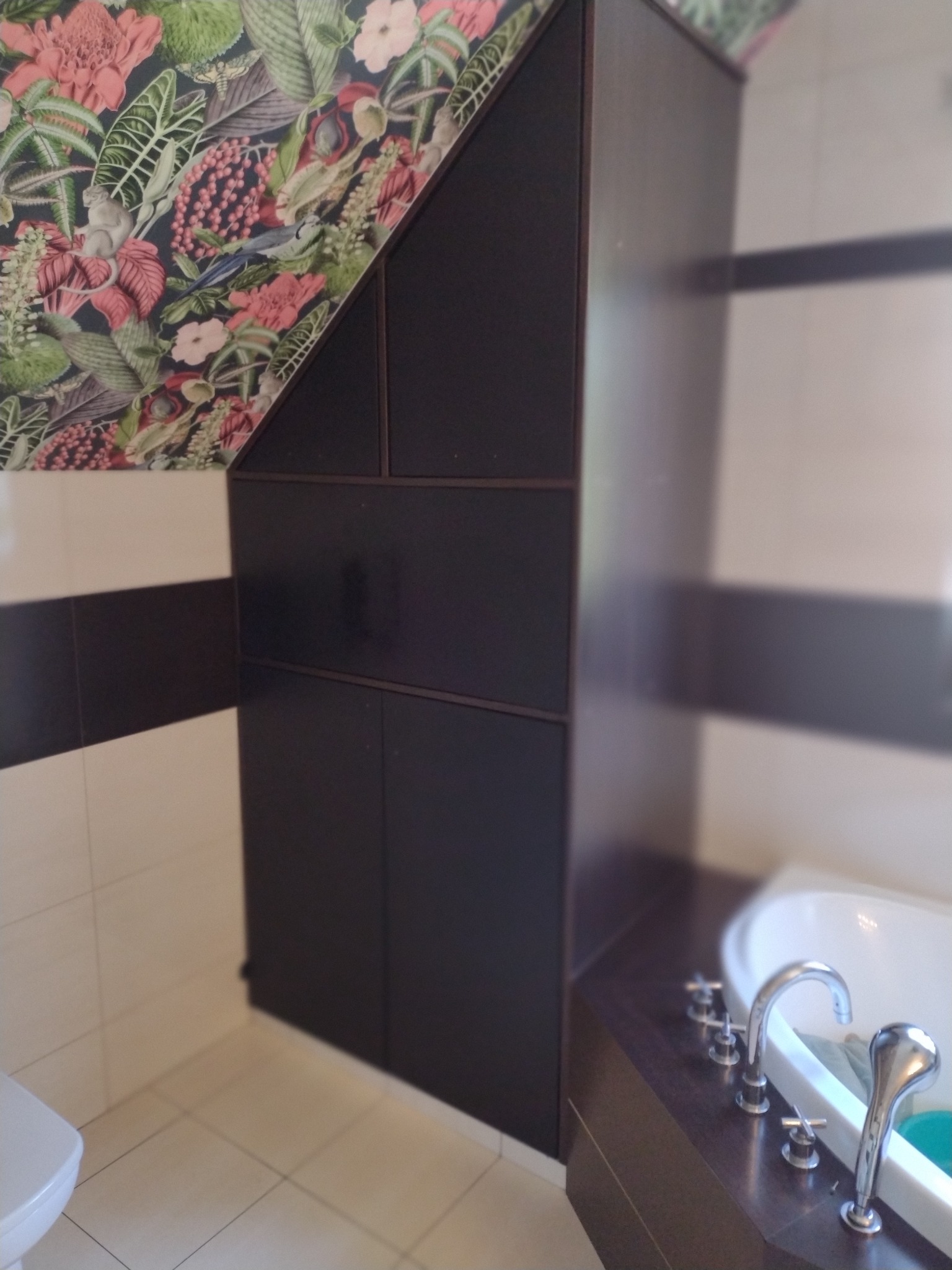 Ciemna zabudowa meblowa w łazience ze skośnym sufitem, ściana pokryta tapetą w kwiaty i zwierzęta, widoczna część wanny i baterii.