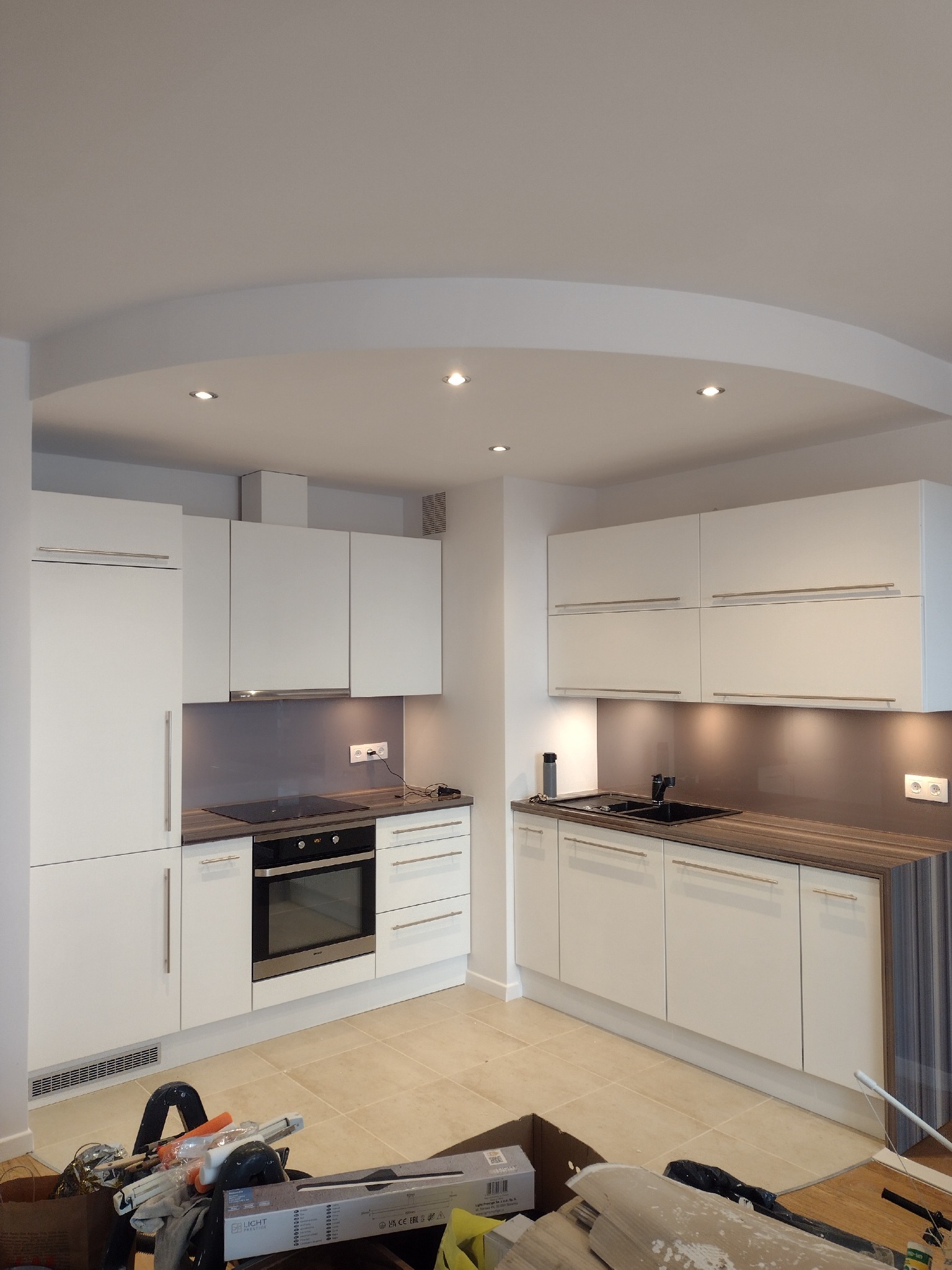 Nowoczesna, jasna kuchnia z białymi szafkami i brązowym blatem w trakcie remontu, widoczne narzędzia i materiały budowlane na podłodze.