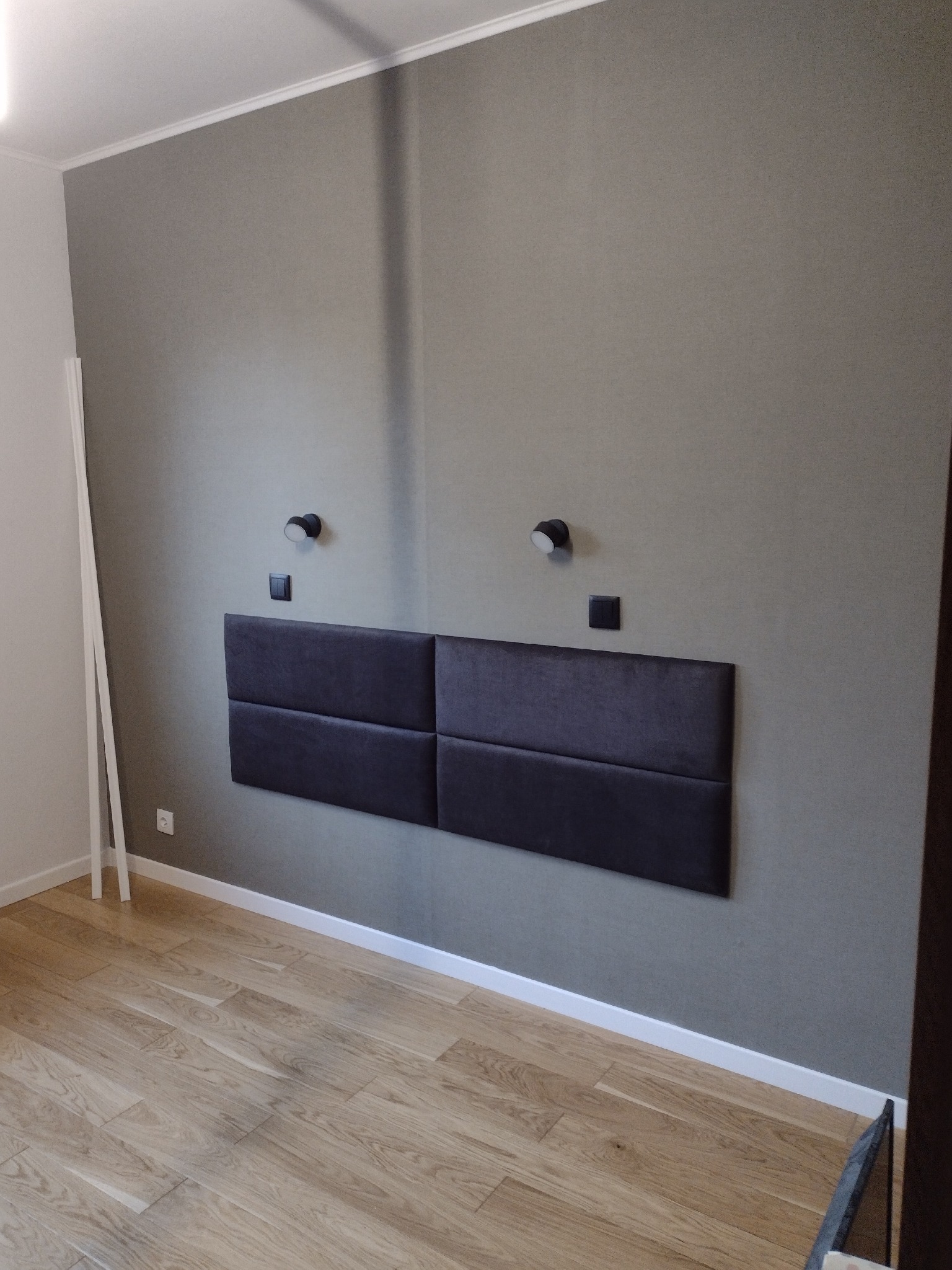 Szara ściana z zamontowanym, ciemnoszarym, tapicerowanym zagłówkiem składającym się z czterech paneli, oświetlonym dwoma czarnymi kinkietami z włącznikami. Jasna drewniana podłoga i białe listwy...