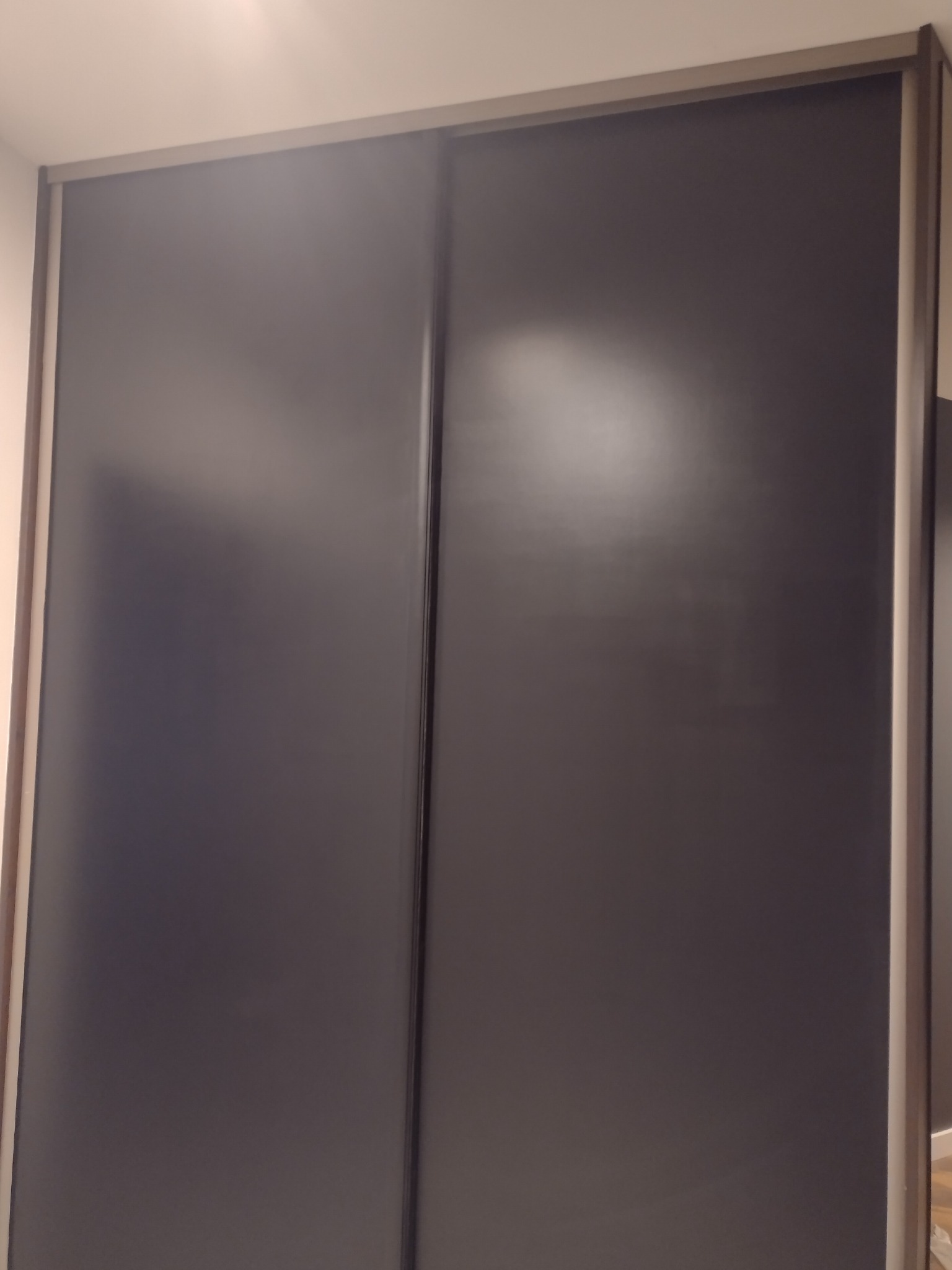 Dwie przesuwne, grafitowe drzwi szafy wnękowej z szarymi obramowaniami, widoczne na tle białej ściany i sufitu.