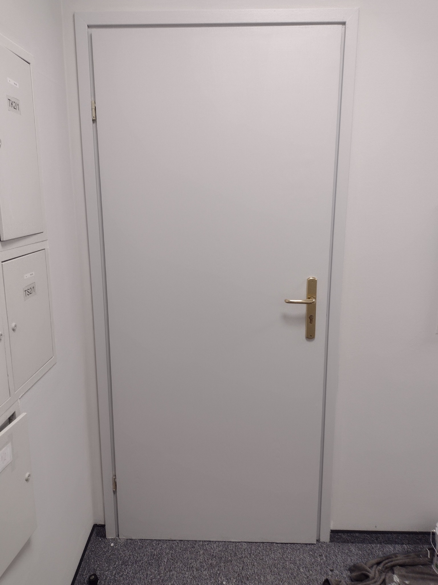 Białe, pomalowane drzwi wewnętrzne z metalową ościeżnicą i złotą klamką w biurze, widoczne szafki elektryczne po lewej stronie.