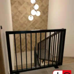 Nowoczesna, czarna balustrada schodowa z pionowymi prętami, widok z góry, w tle ściana z geometrycznym wzorem i wisząca lampa z kulistymi kloszami, w prawym dolnym rogu widoczne logo firmy.