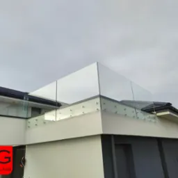 Nowoczesna, bezramowa konstrukcja ze szkła na balkonie, mocowana punktowo do ściany budynku o białej elewacji, widoczny fragment dachu z czarnej dachówki i logo firmy w lewym dolnym rogu.