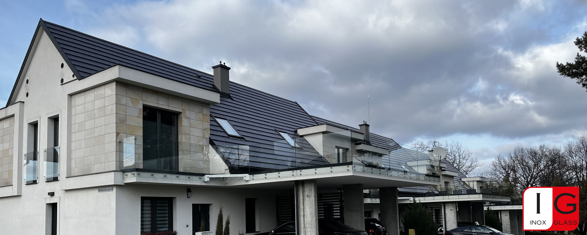 Nowoczesny dom z jasną elewacją i ciemnym dachem, wyposażony w szklane balustrady balkonowe i tarasowe, widoczne z zewnątrz. W tle pochmurne niebo i drzewa.