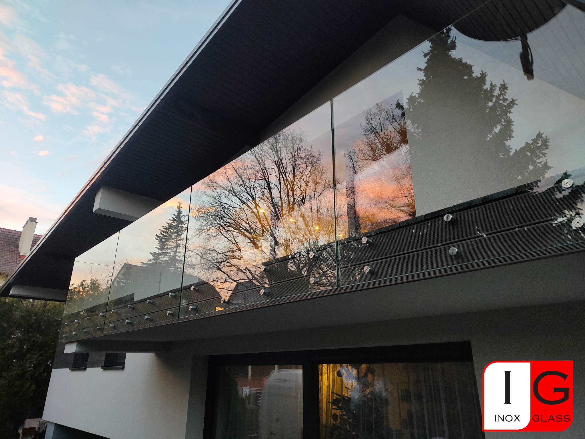 Nowoczesna balustrada ze szkła na balkonie, mocowana punktowo, z widocznym odbiciem drzew i zachodzącego słońca.