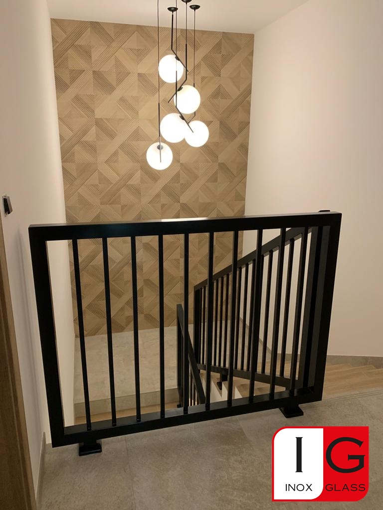 Nowoczesna, czarna balustrada schodowa z pionowymi prętami, widok z góry, w tle ściana z geometrycznym wzorem i wisząca lampa z kulistymi kloszami, w prawym dolnym rogu widoczne logo firmy.