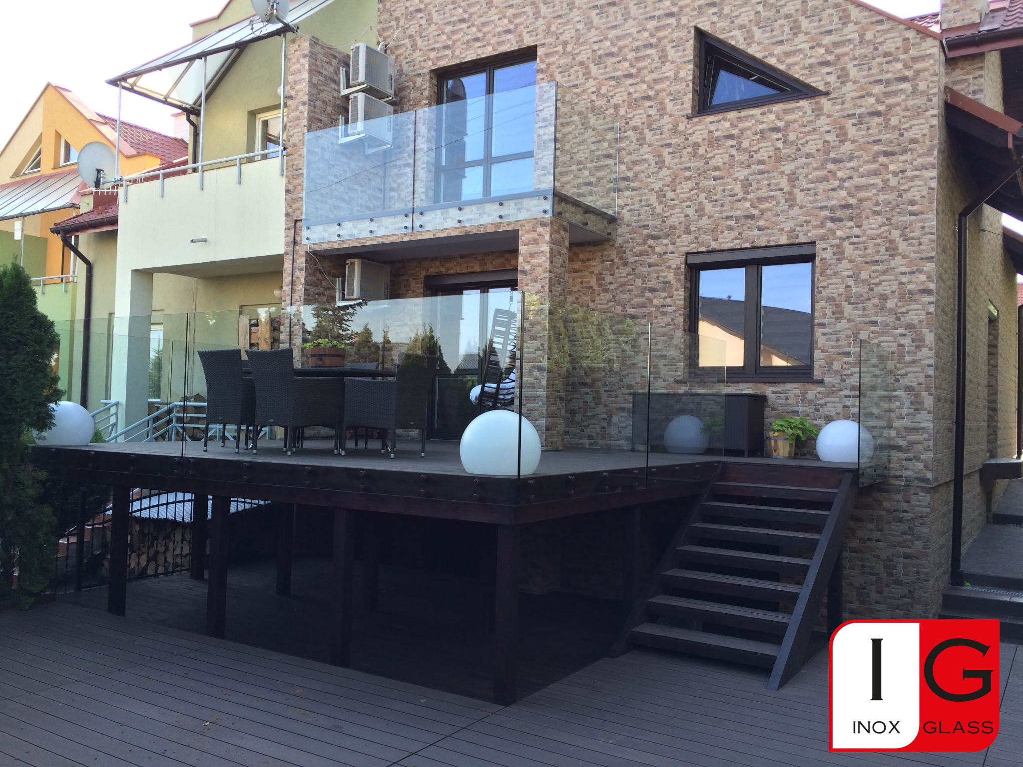 Nowoczesny dom z tarasem zabezpieczonym szklaną balustradą, widok z zewnątrz, z widocznym logo firmy na dole po prawej.