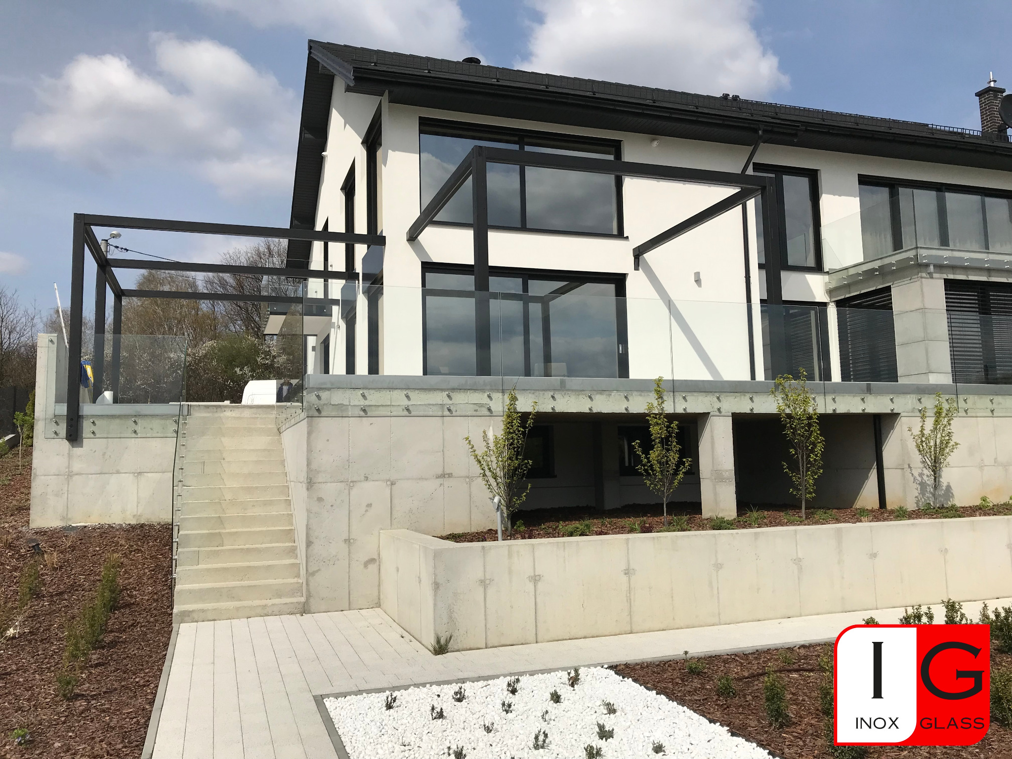 Nowoczesny dom z białymi ścianami i czarnymi ramami okiennymi, wyposażony w szklane balustrady na tarasie i balkonie, z widocznym logo firmy Inox Glass w prawym dolnym rogu.
