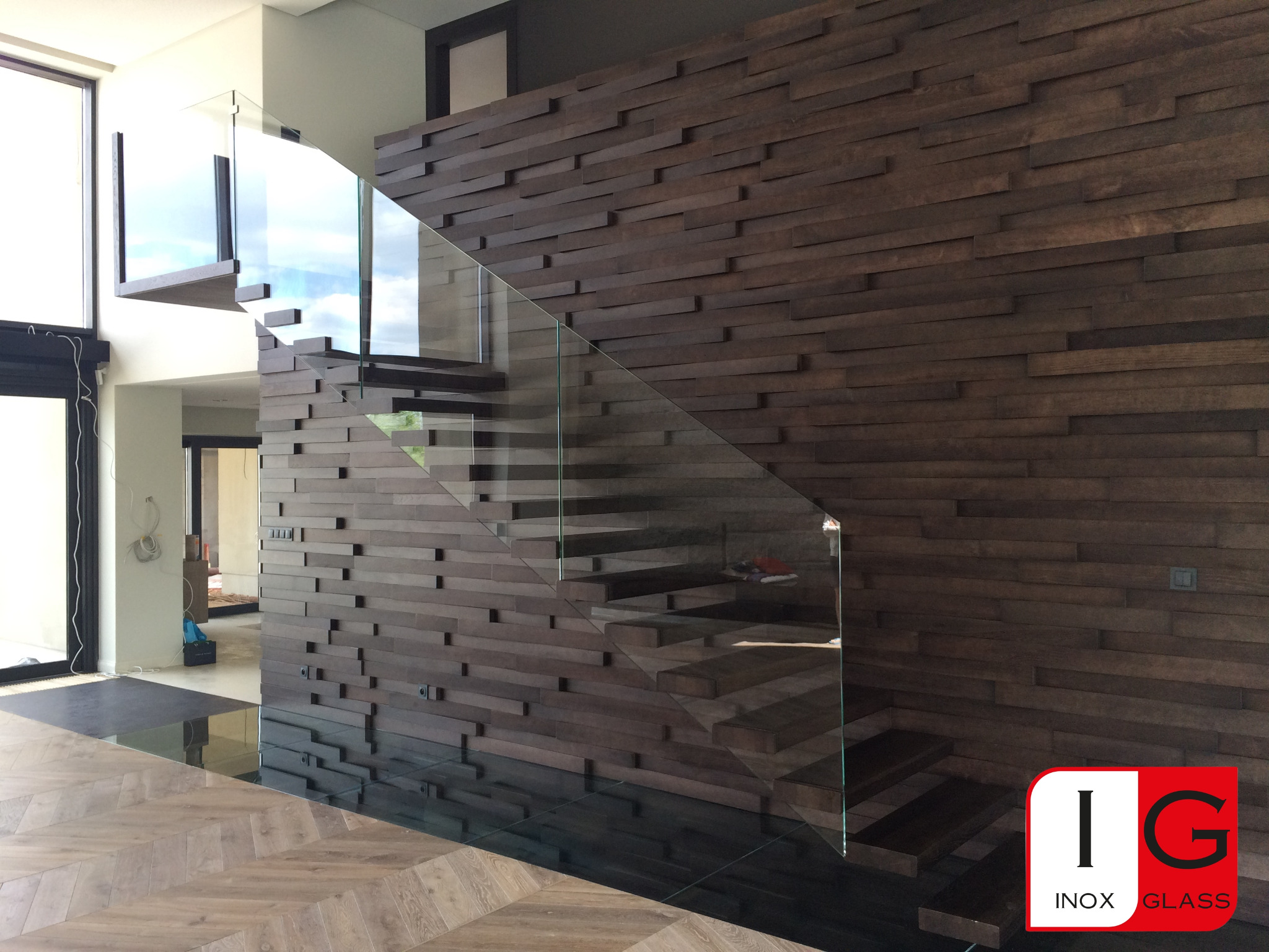 Nowoczesne schody z drewnianymi stopniami i szklaną balustradą, widoczne na tle ściany z dekoracyjnych drewnianych paneli, z logo firmy w prawym dolnym rogu.