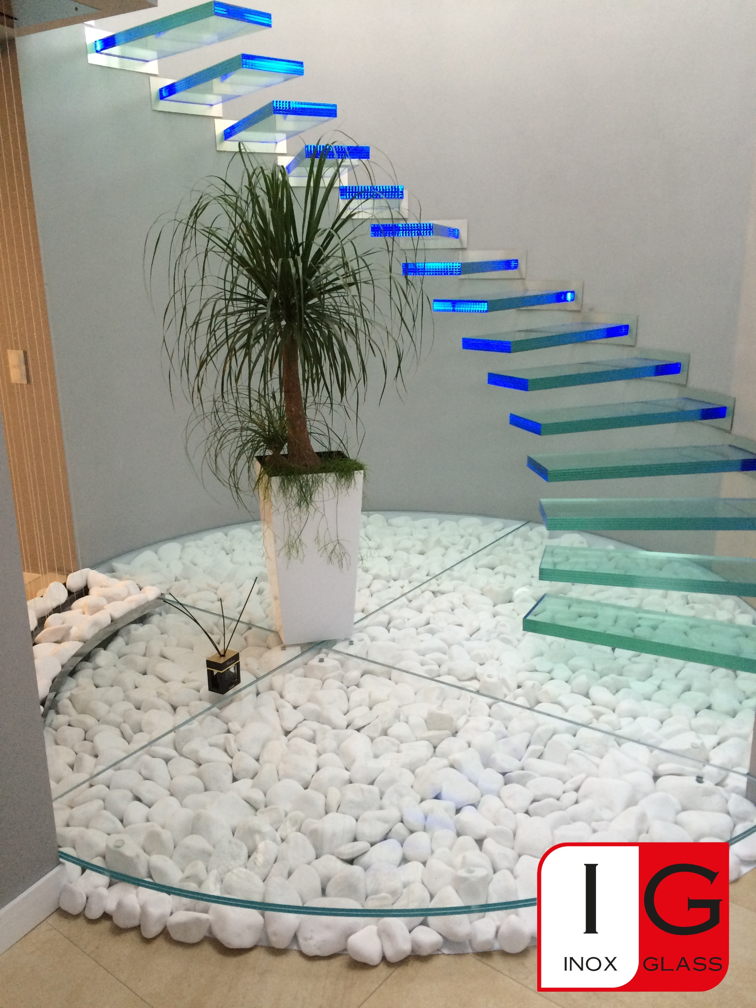 Szklane schody spiralne z niebieskim podświetleniem LED, osadzone na szklanej platformie wypełnionej białymi kamieniami, obok donica z rośliną, widoczne logo firmy Inox Glass w rogu.