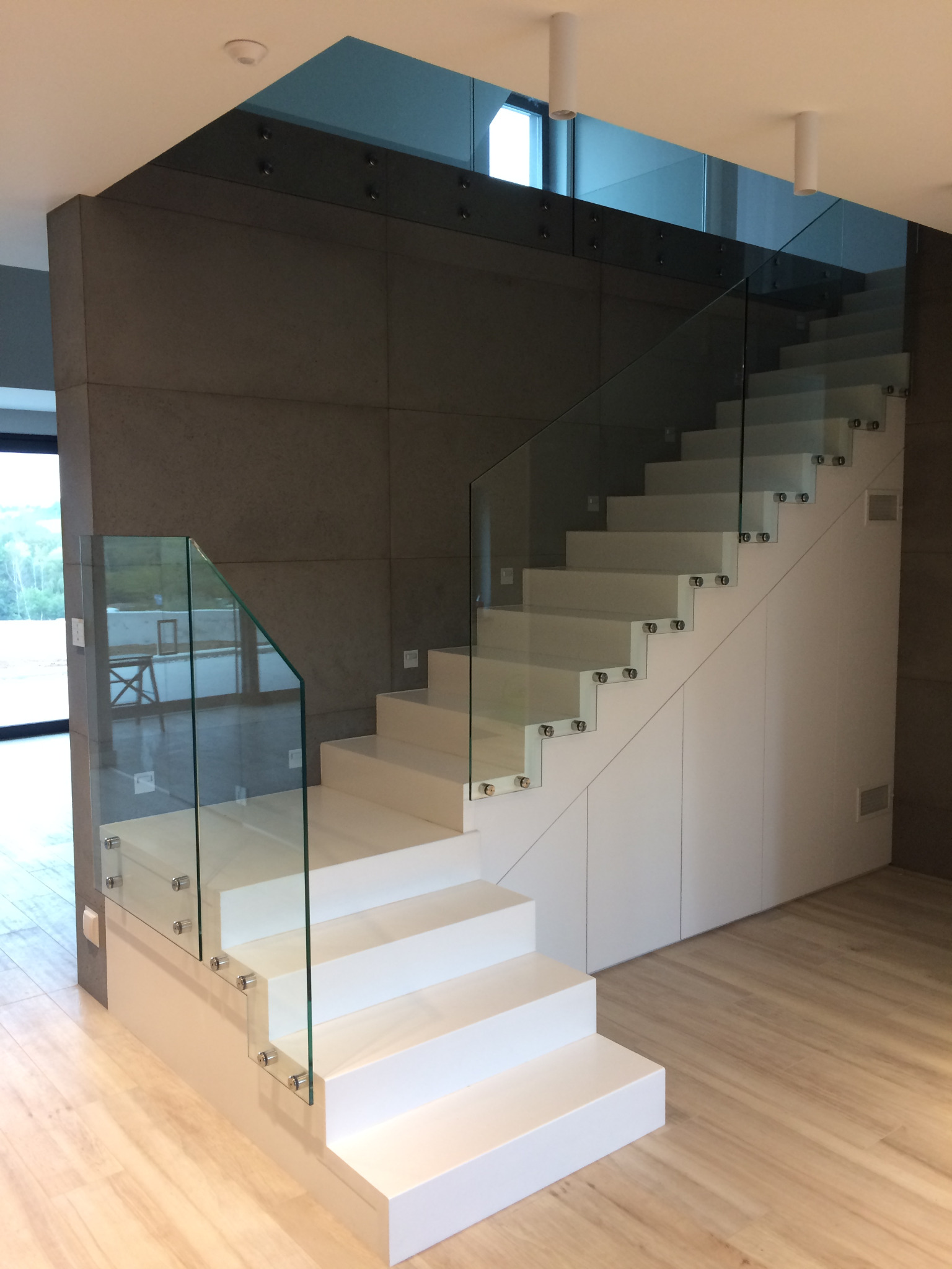 Nowoczesne schody z białymi stopniami i szklaną balustradą mocowaną punktowo, widok na drewnianą podłogę i szarą ścianę w tle.